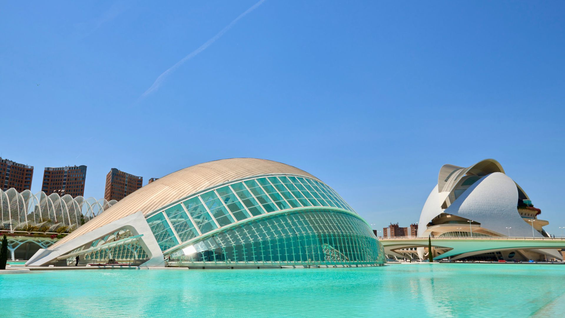 Futuristic glass and white buildings beside a blue pool under a clear sky.