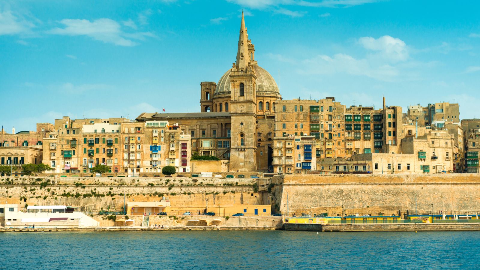 Historic waterfront buildings and a large domed church in Valletta, Malta, with blue sky and water in front.