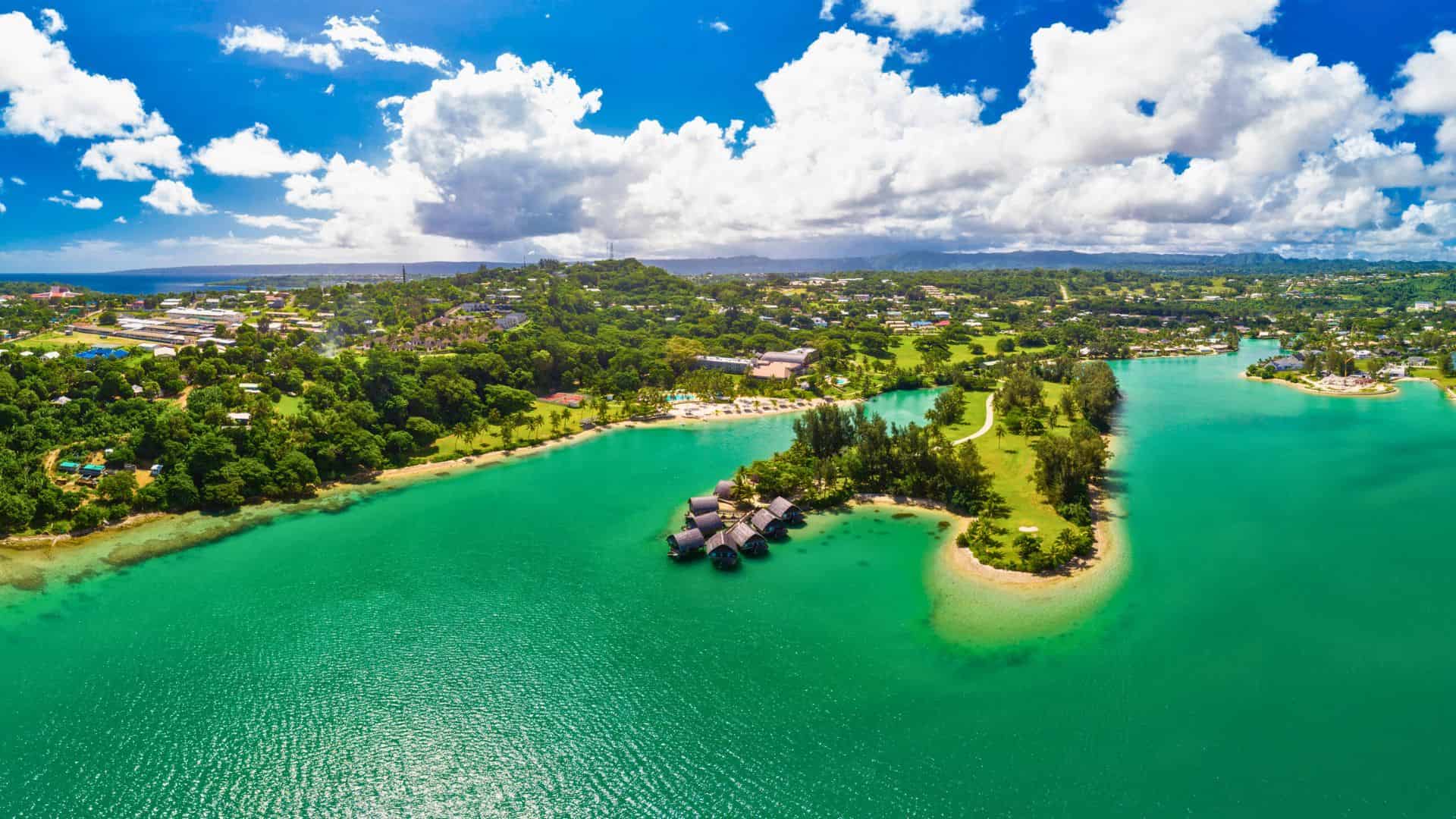 Aerial view of a tropical island with turquoise water, lush greenery, and scattered buildings under a partly cloudy sky.