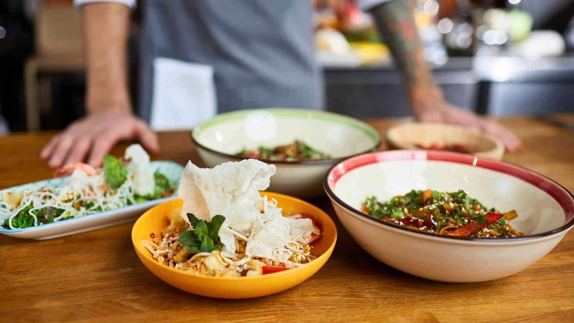 Four colorful bowls of assorted food on a wooden table with a chef standing in the background.