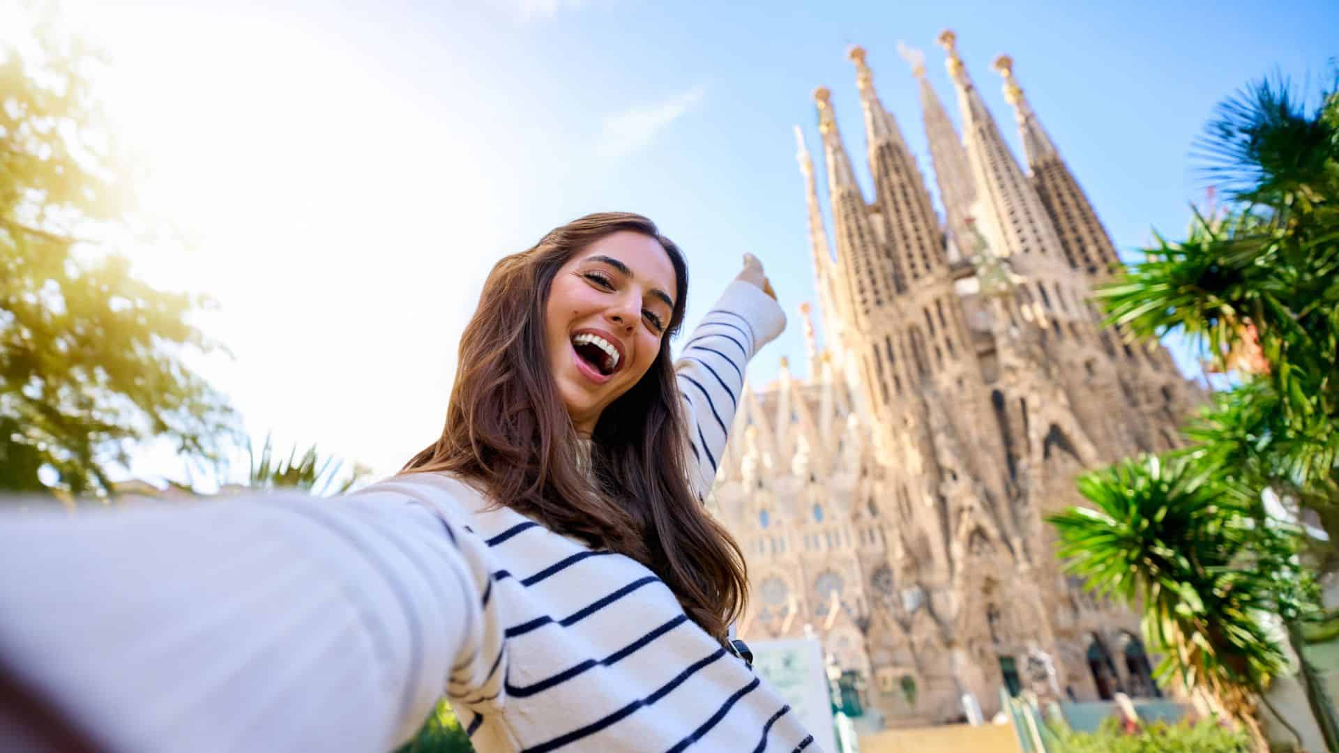Woman taking a selfie and smiling in front of La Sagrada Familia in Barcelona on a sunny day.