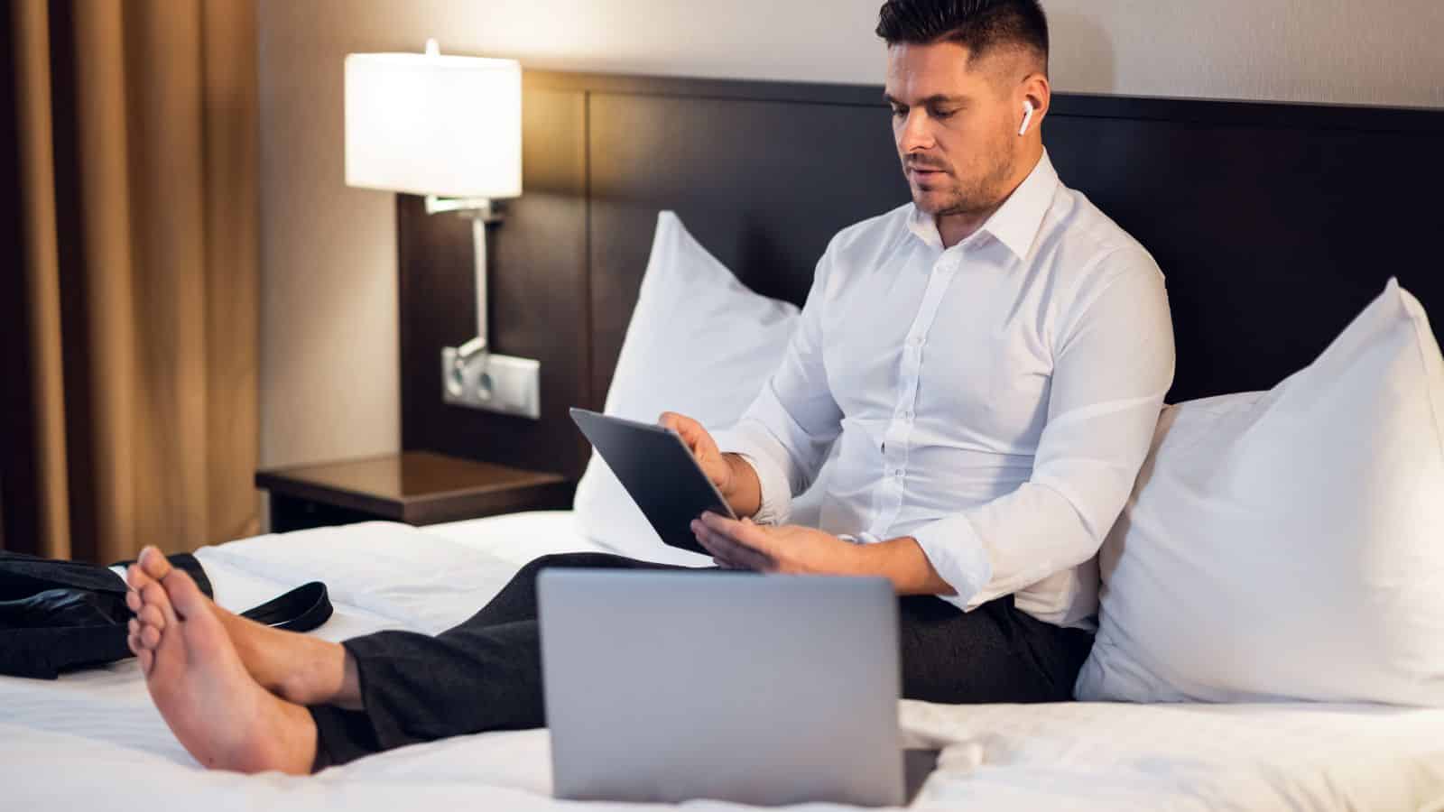 Man in dress shirt sits on hotel bed using tablet, with a laptop nearby and wireless earphones in his ears&mdash;enjoying travel advice and hotel booking tips during his stay.