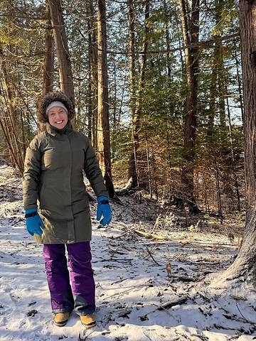 Bundled up for winter travel, a person in a coat, gloves, and boots stands on a snowy forest path as sunlight filters through the trees.