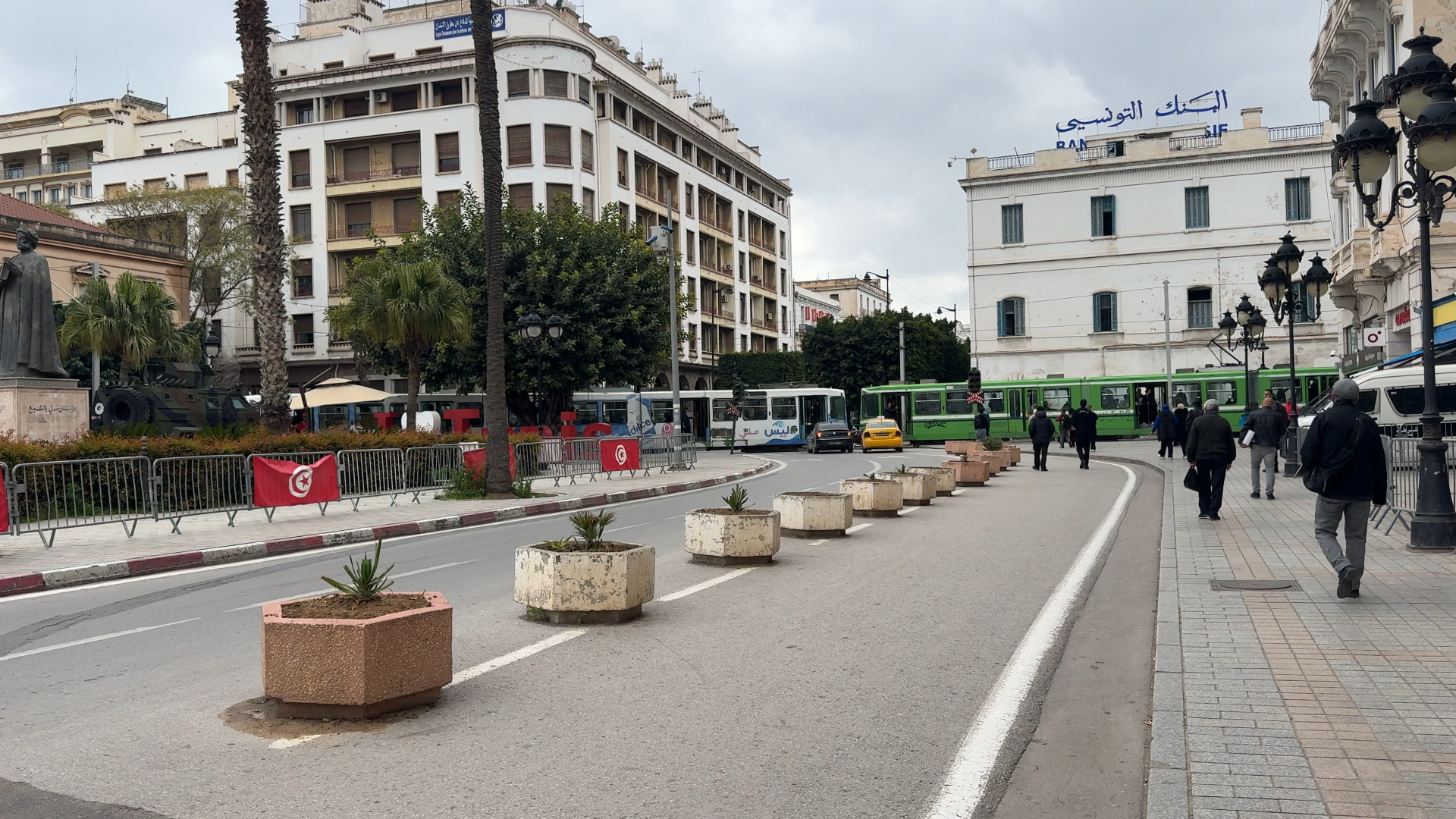 A lively street in a city, with potted plants dividing lanes—people walking, buses passing, and buildings rising behind—captures the vibrant atmosphere perfect for a Tunisia itinerary 1 day experience.