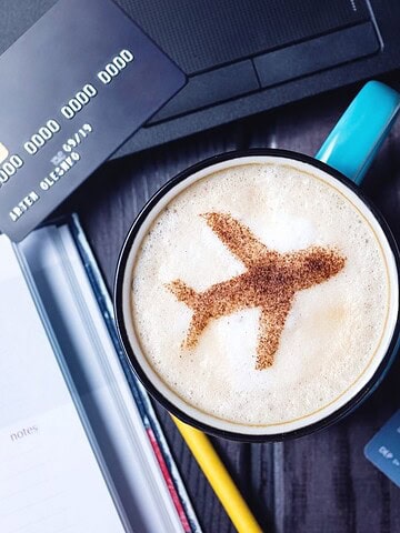 A coffee cup with an airplane design in foam, beside flight tickets, a credit card, and a laptop.