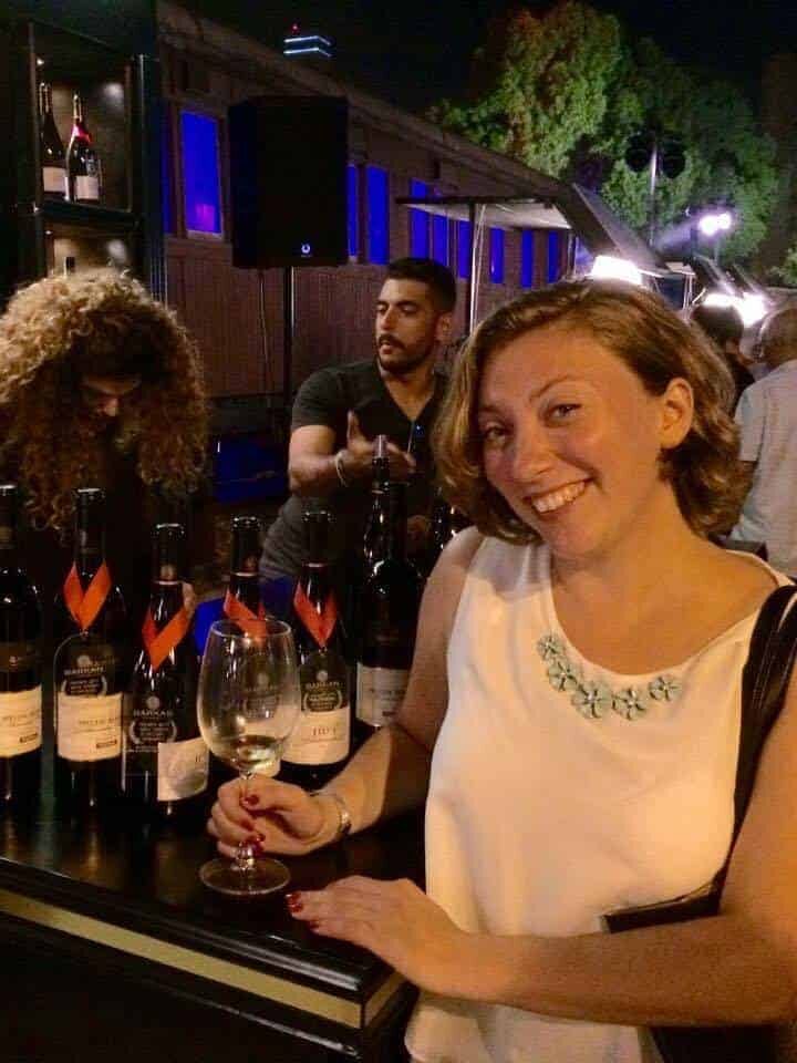Woman smiling with a glass of wine at a nighttime wine tasting event, bottles displayed on the table.
