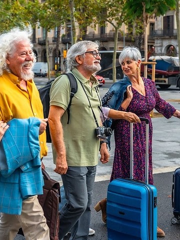 Four smiling older adults with suitcases walk together on a city street lined with trees and buildings.