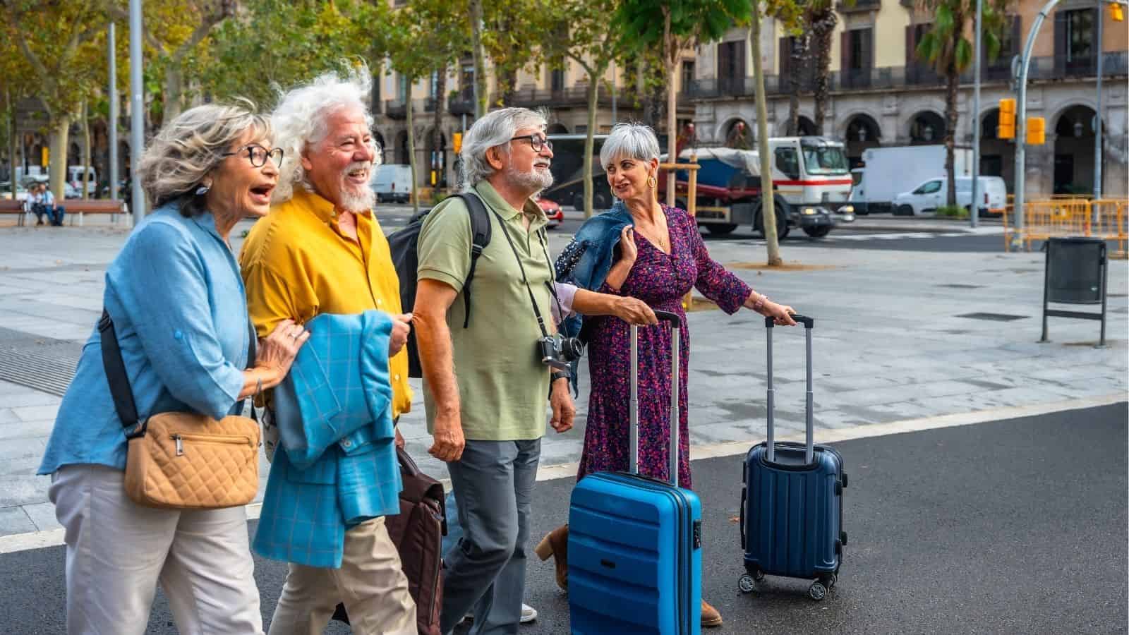 Four smiling older adults with suitcases walk together on a city street lined with trees and buildings.