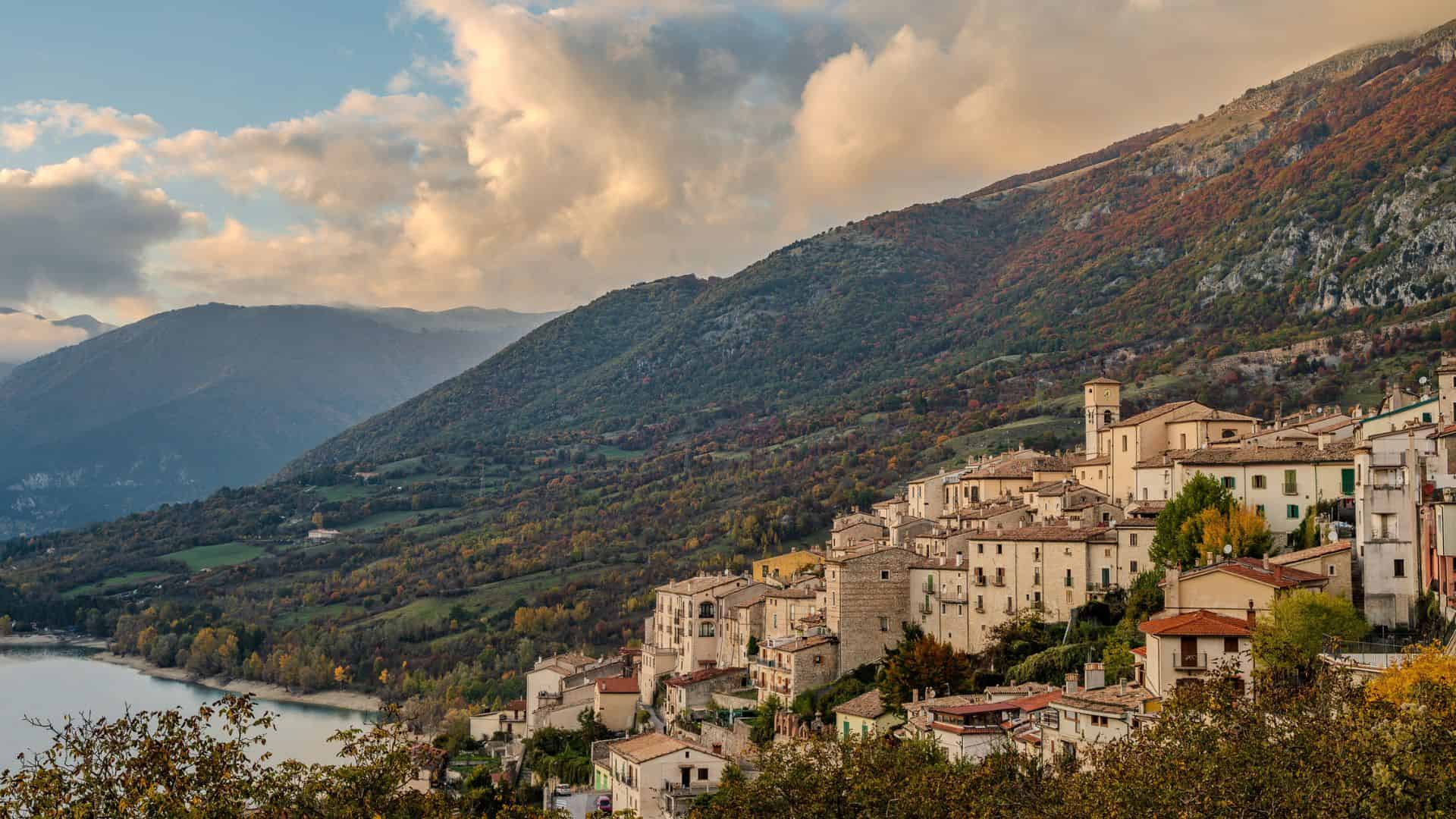 Hilltop village with stone buildings overlooking a lake and forested mountains under a cloudy sky.