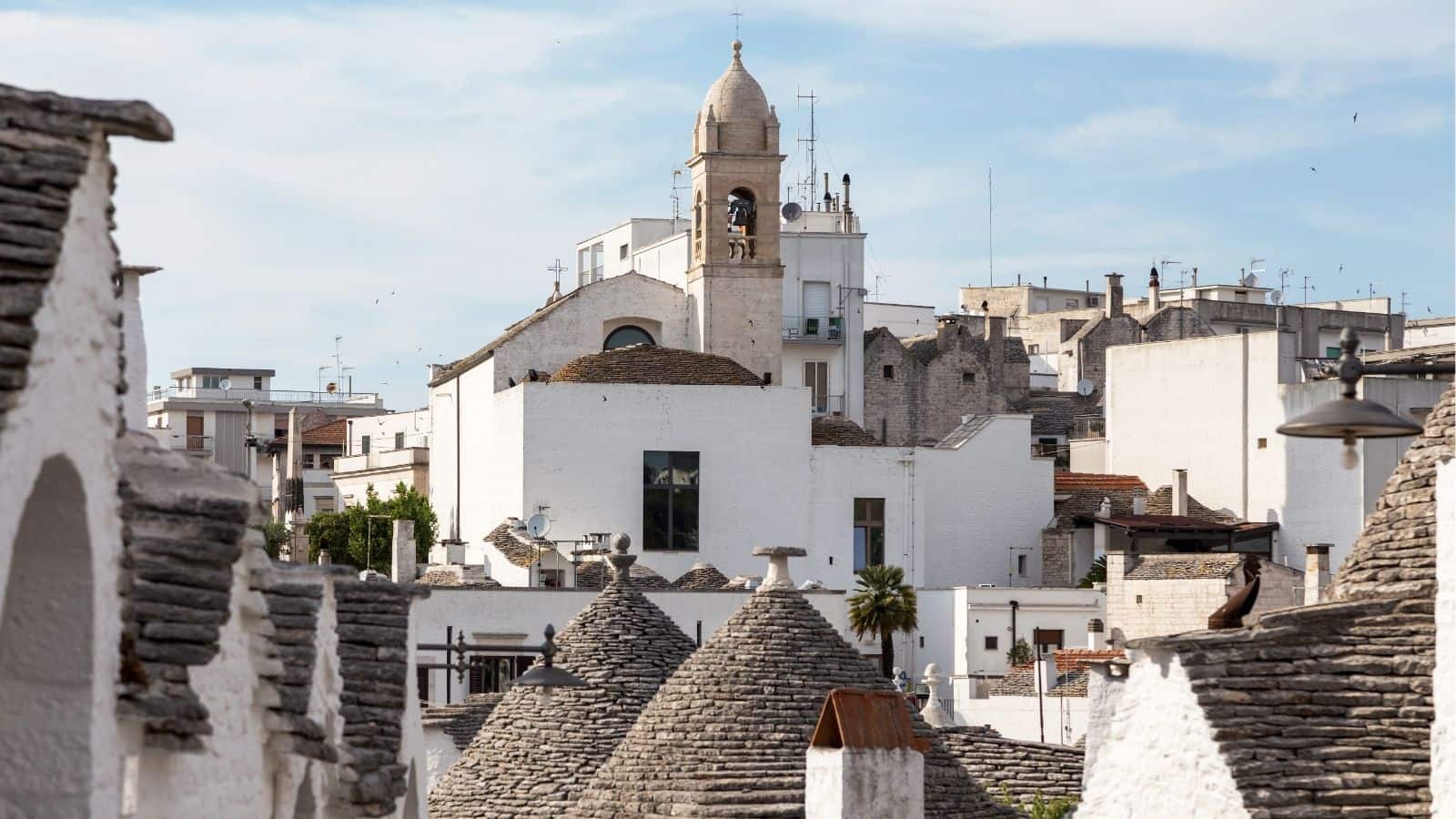 White stone buildings and conical roofs with a church tower in the background under a blue sky.