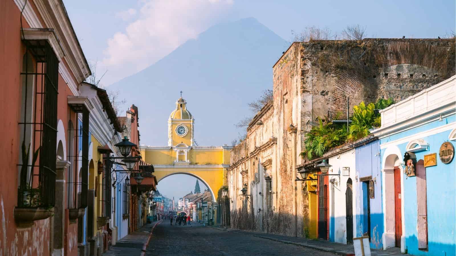 A cobblestone street leads to a yellow arch with a clock, with a volcano visible in the background.