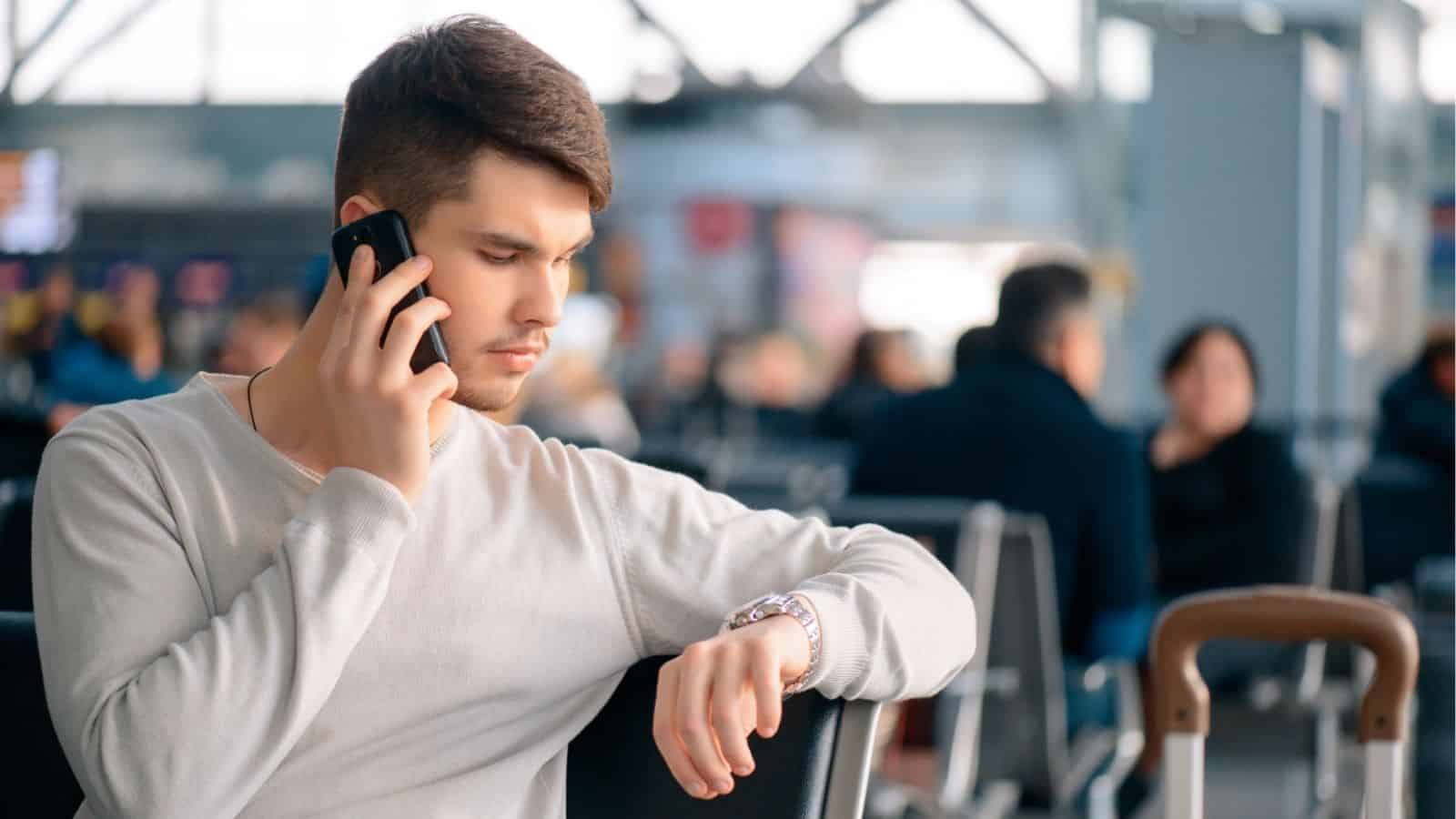 A man at an airport checks his watch while talking on the phone, with blurred travelers in the background.