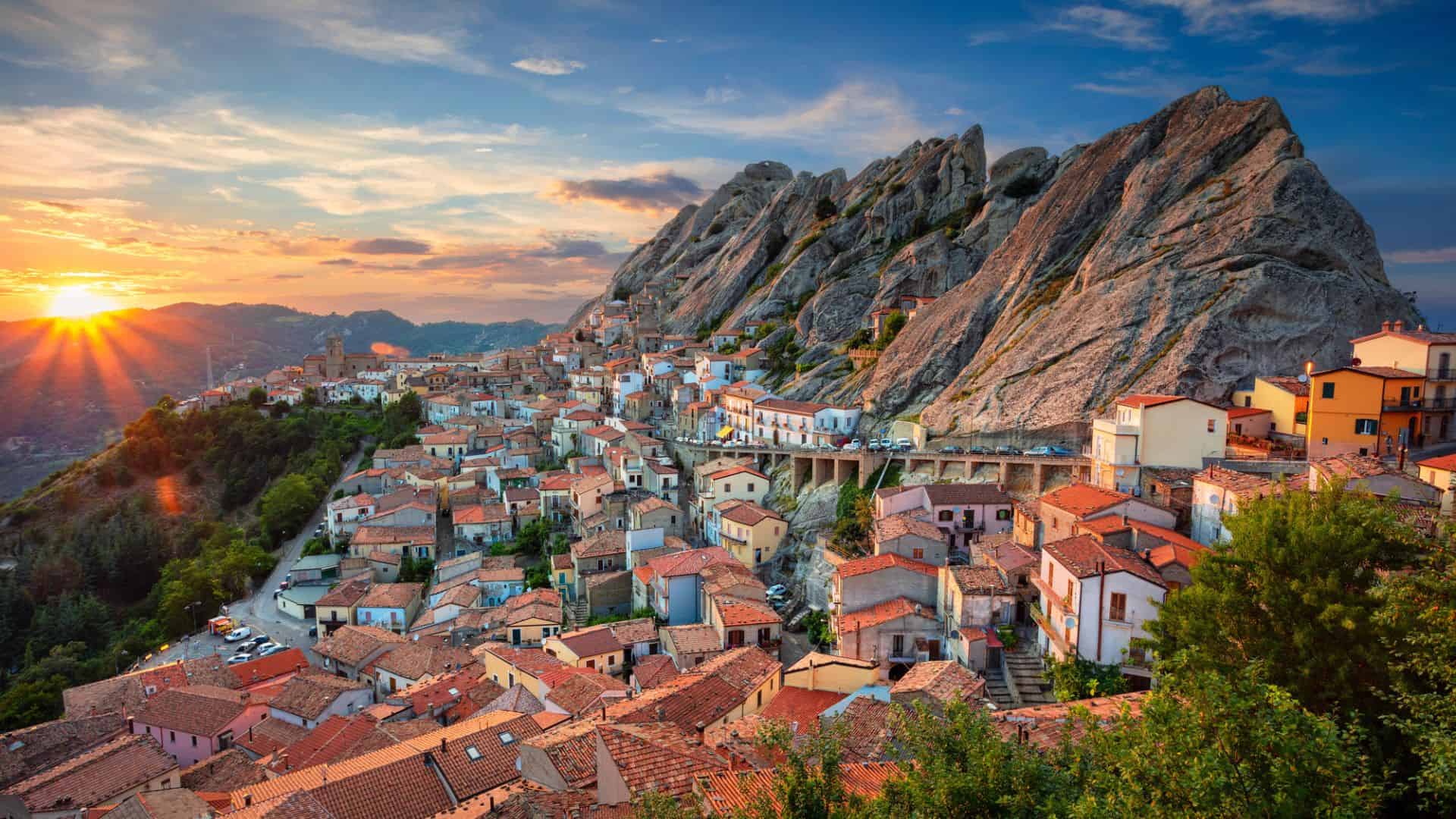 Colorful hillside village with terracotta roofs at sunset, nestled beside dramatic rocky cliffs and lush greenery.