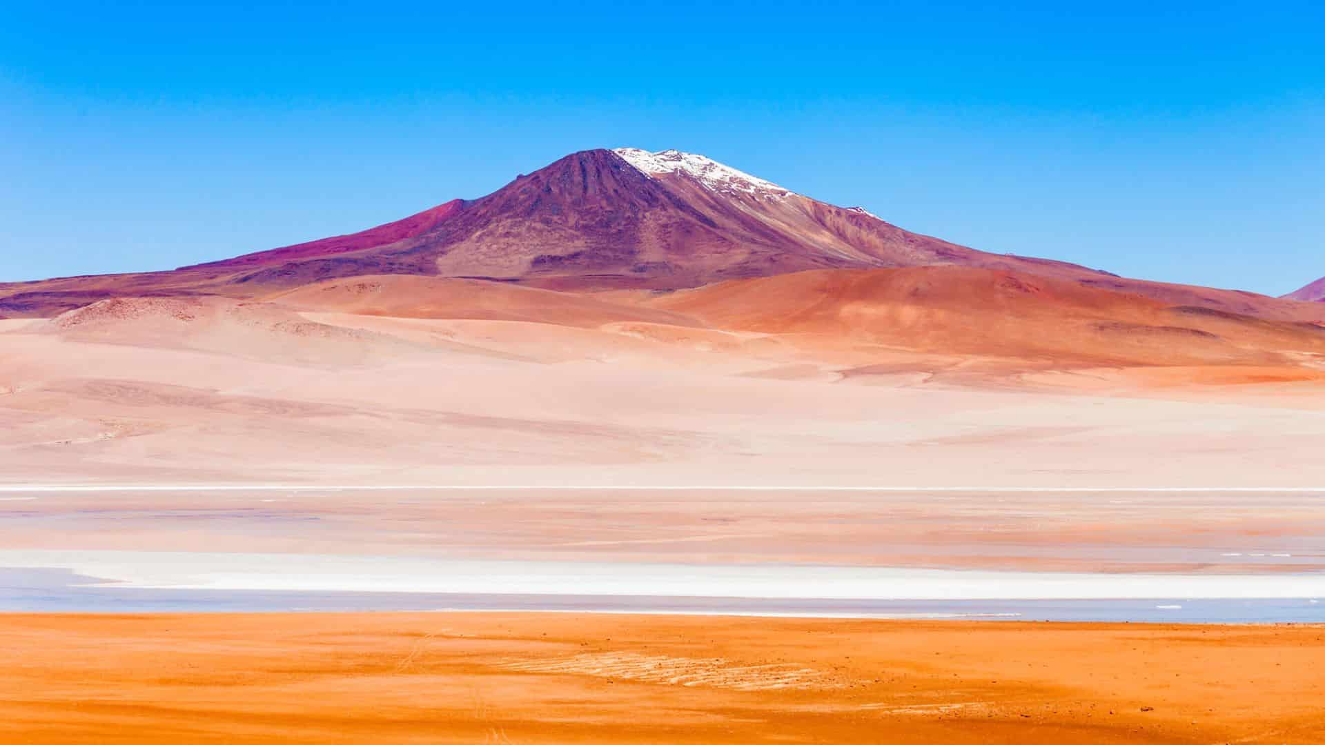 Snow-capped mountain with colorful, barren desert terrain under a clear blue sky.