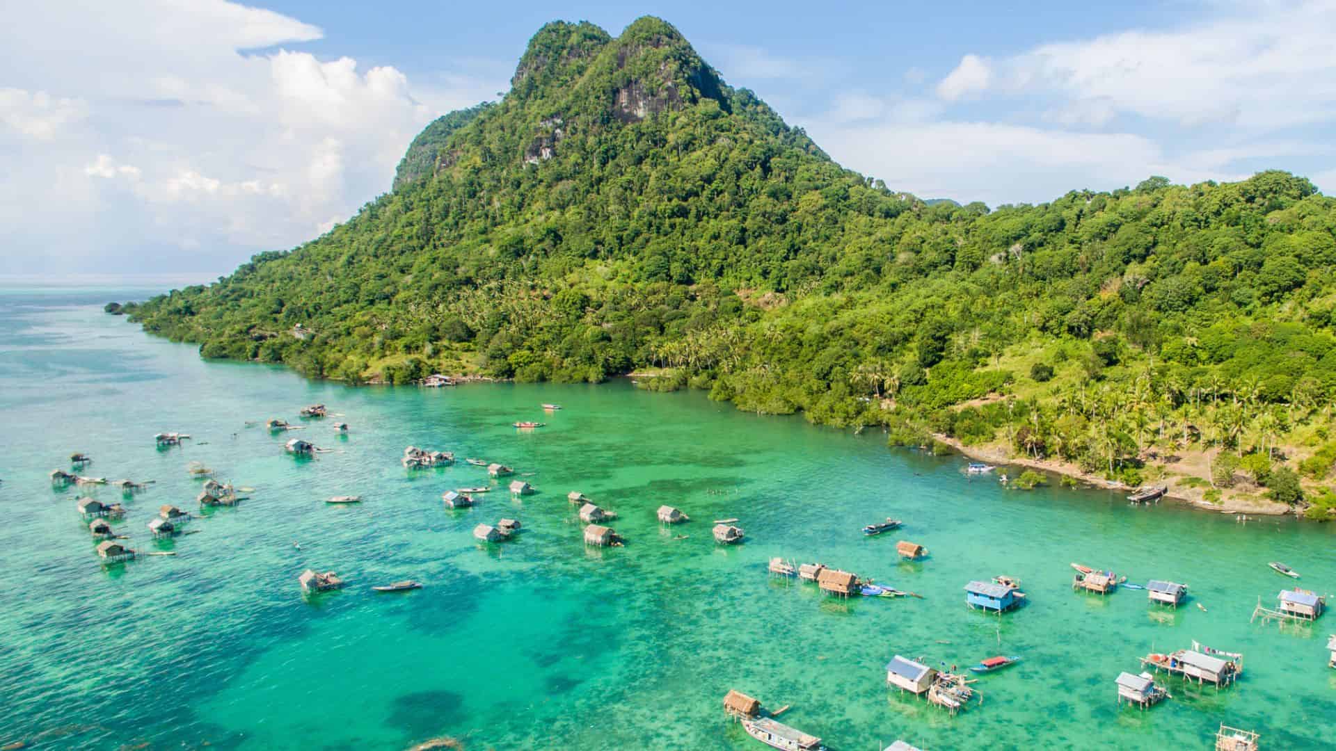 Aerial view of turquoise water, stilt houses, and a lush green hill on a tropical island.