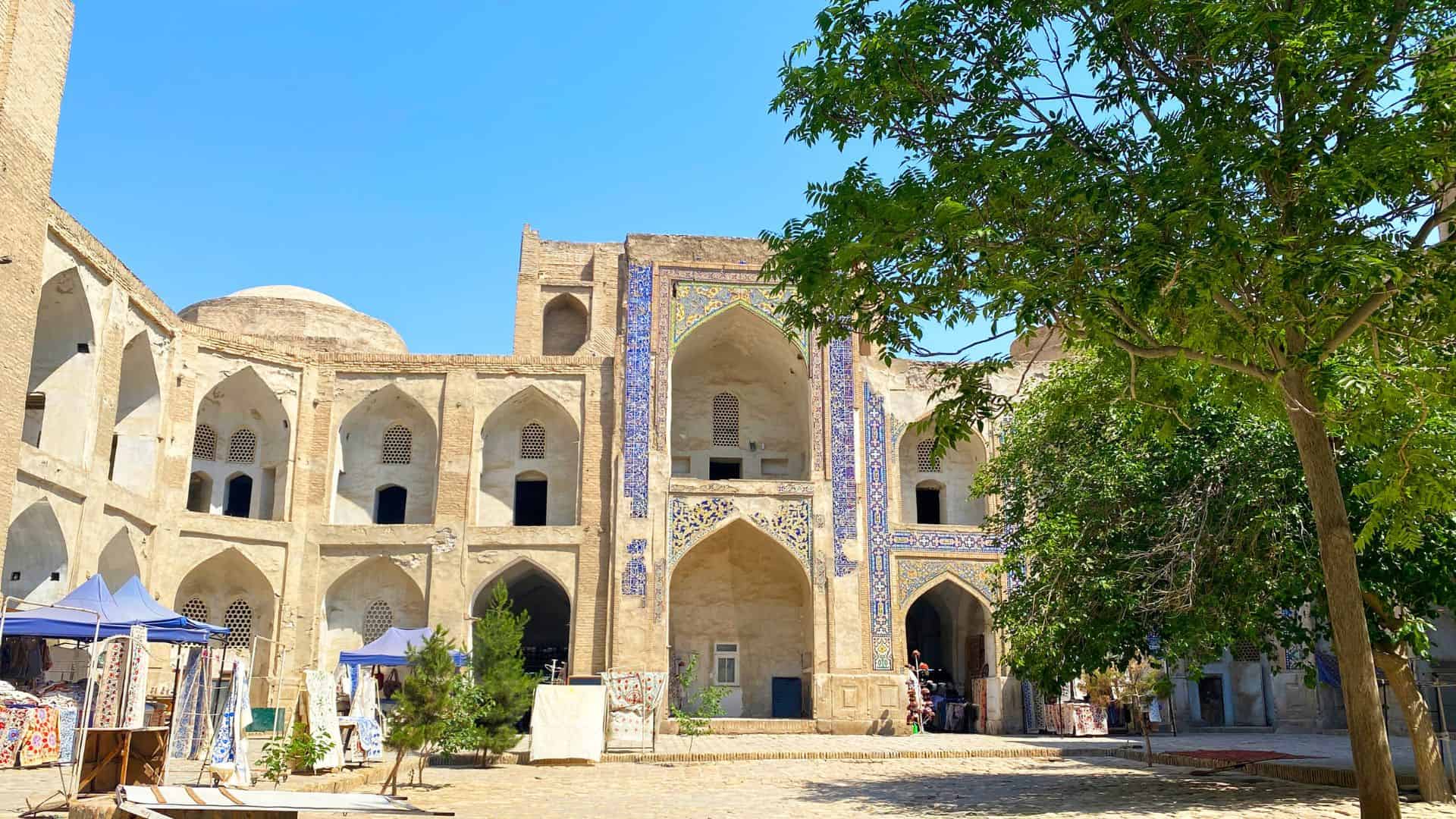 Historic building with arched doorways, mosaic tiles, and trees in a sunny courtyard.