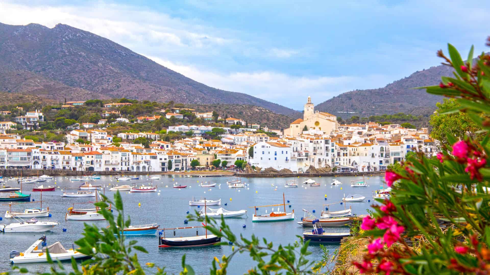Colorful boats on a calm bay, with a white hillside town and mountains in the background, framed by flowers.
