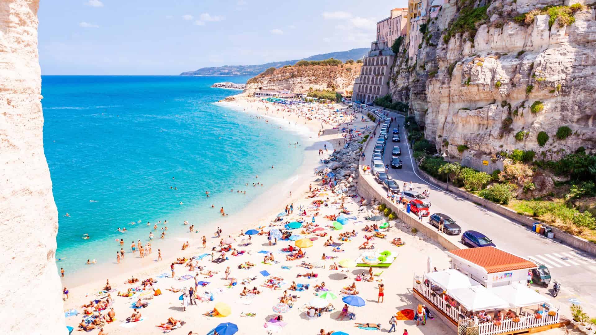 Crowded sandy beach with colorful umbrellas, clear blue sea, and cliffs with buildings and a road alongside.