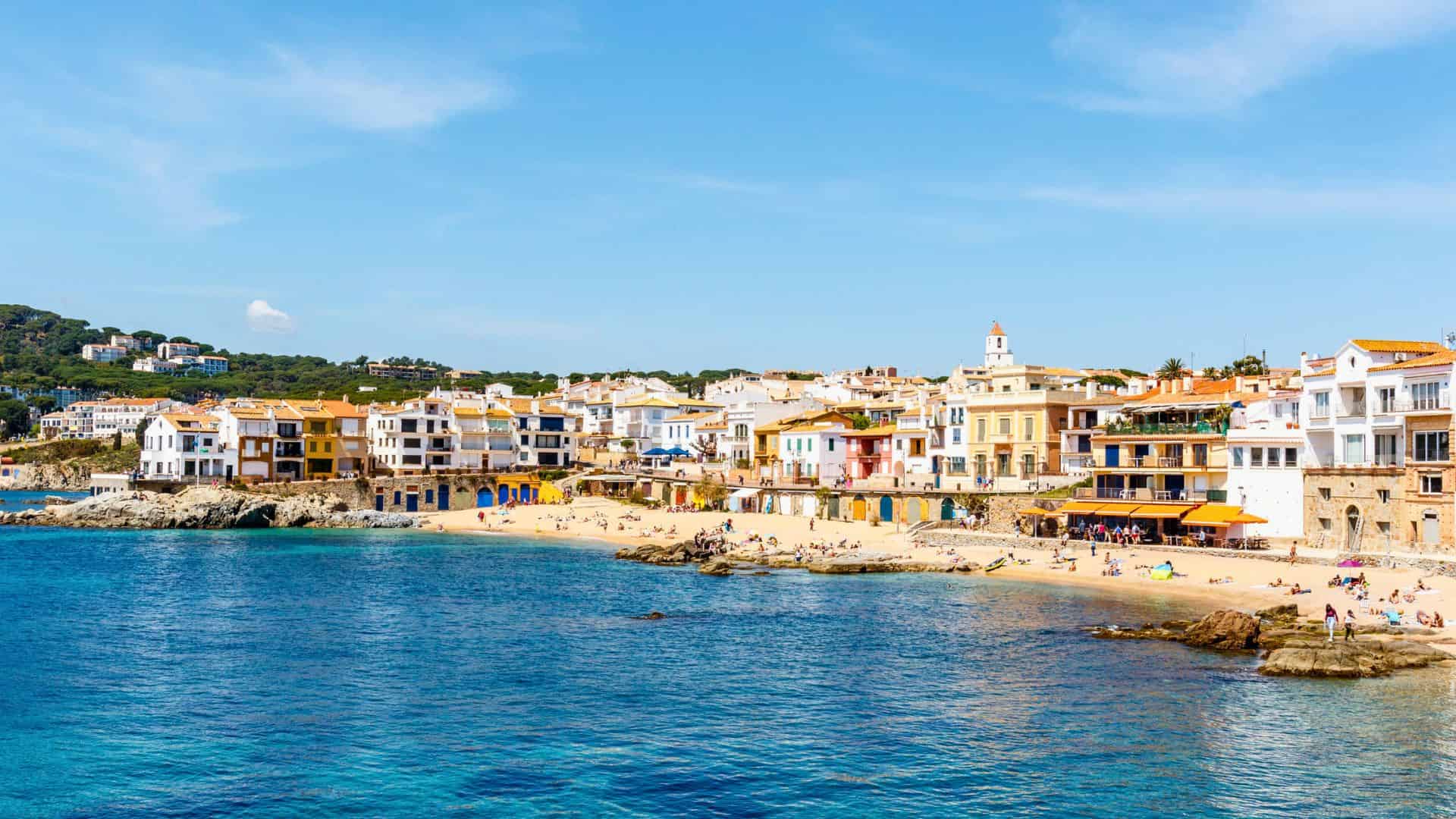 Colorful seaside buildings and people relaxing on a sandy beach by clear blue water under a sunny sky.