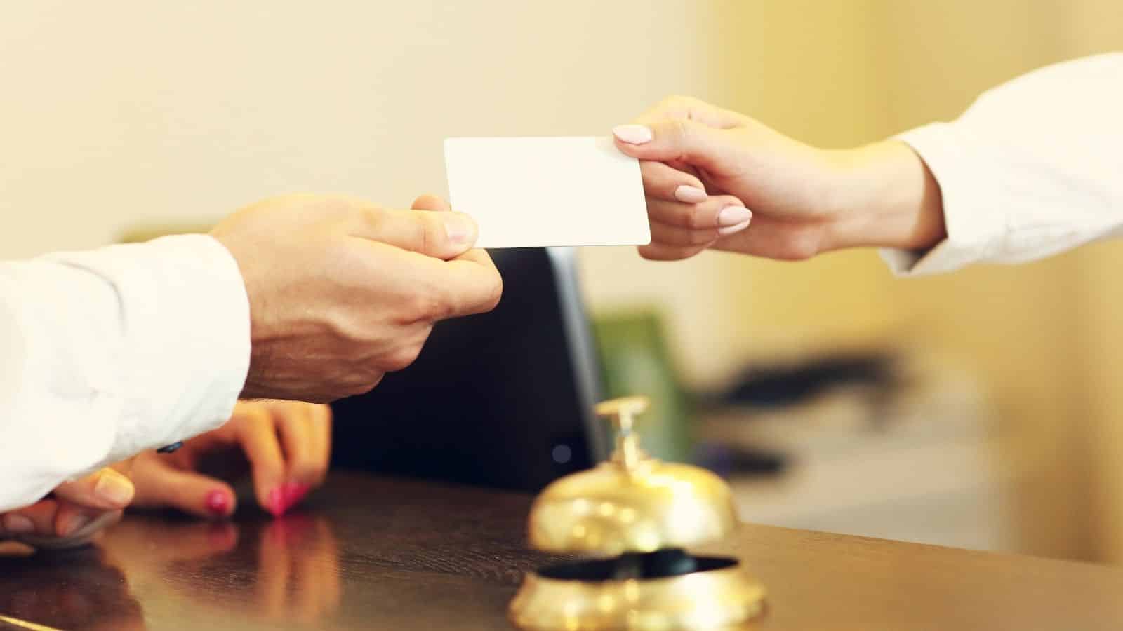 A person hands a key card to another at a hotel front desk with a service bell in the foreground.