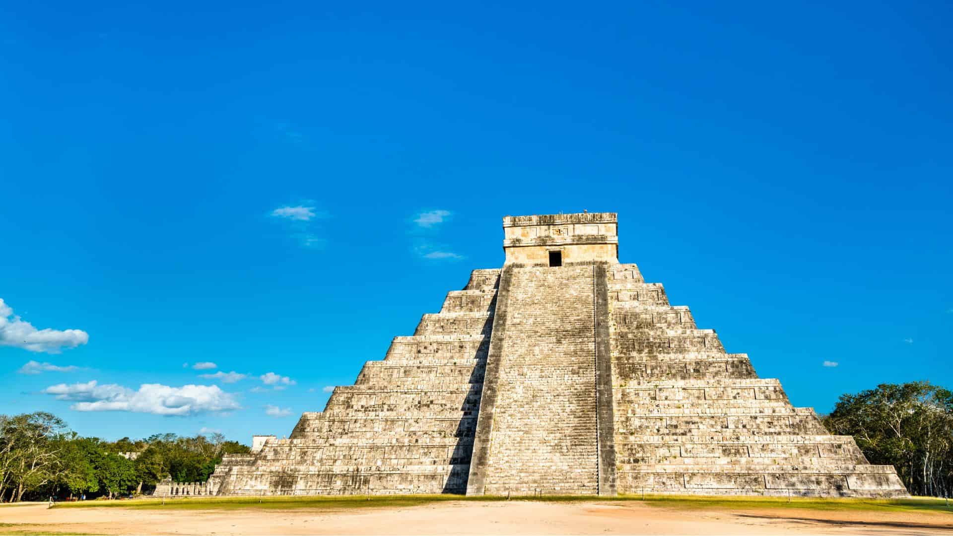 Ancient stone pyramid of Chichen Itza under a clear blue sky with green trees in the background.