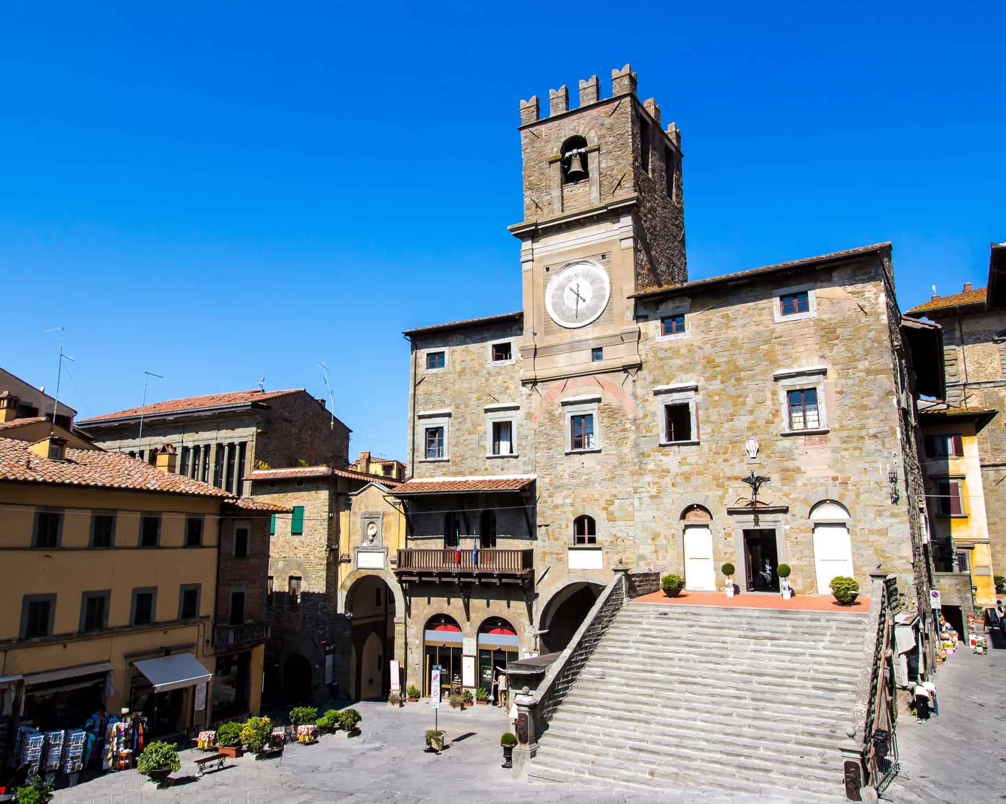 Historic stone building with a clock tower and wide staircase in a sunny Italian town square, reminiscent of the charming architecture found in Tuscany villages.