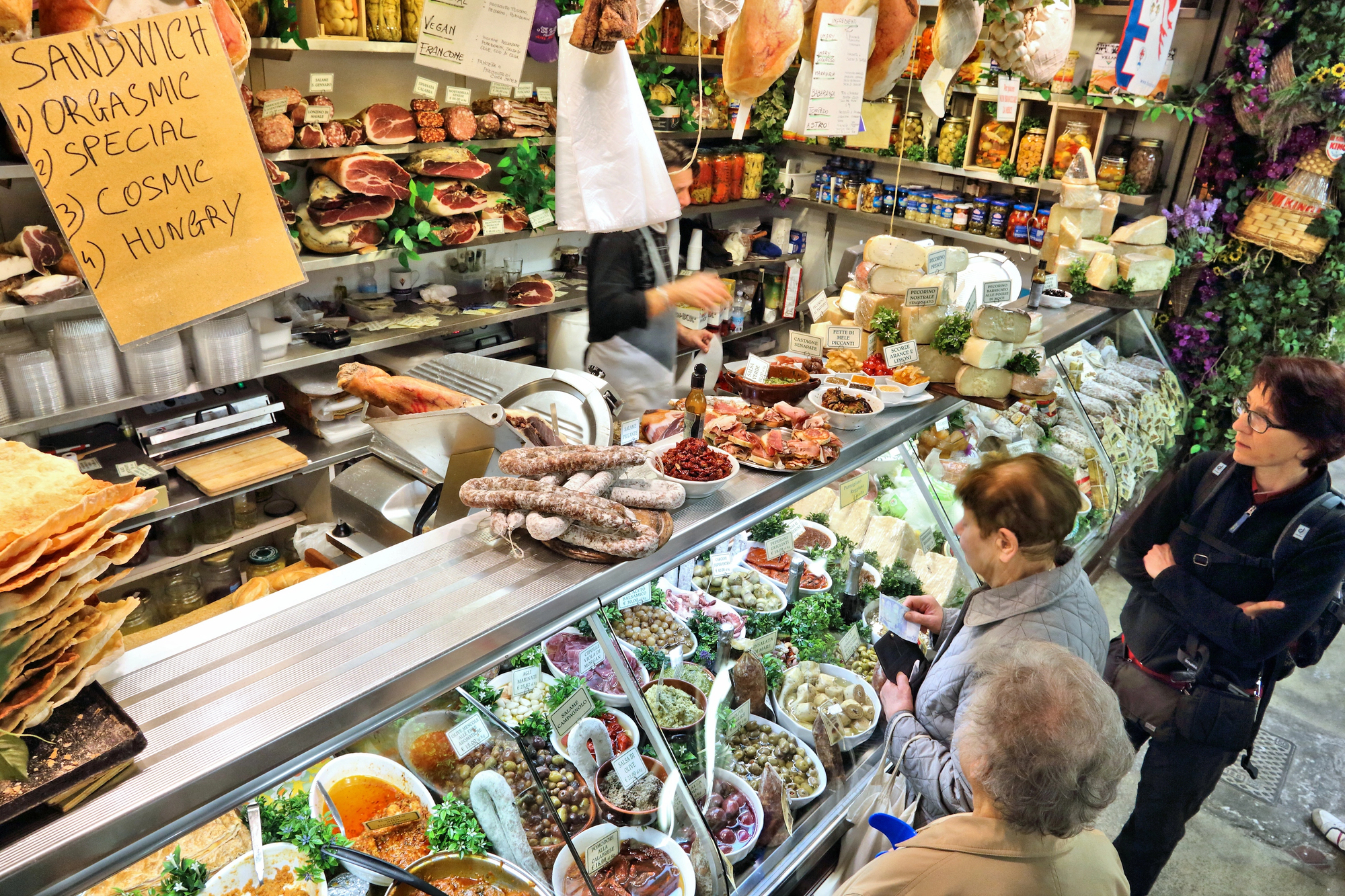 Three people order at a deli counter filled with meats, cheeses, and salads&mdash;hanging meats above evoke the charm of Florence food markets.