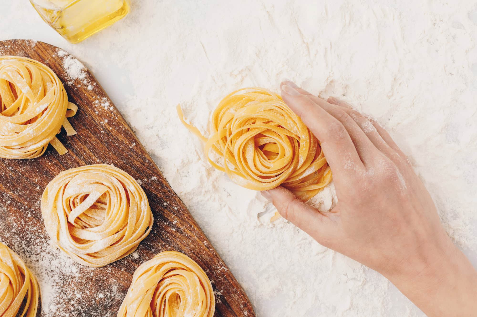 A hand shapes fresh tagliatelle nests on a floured surface, evoking the rustic charm of Tuscany, with more pasta resting on a wooden board nearby.