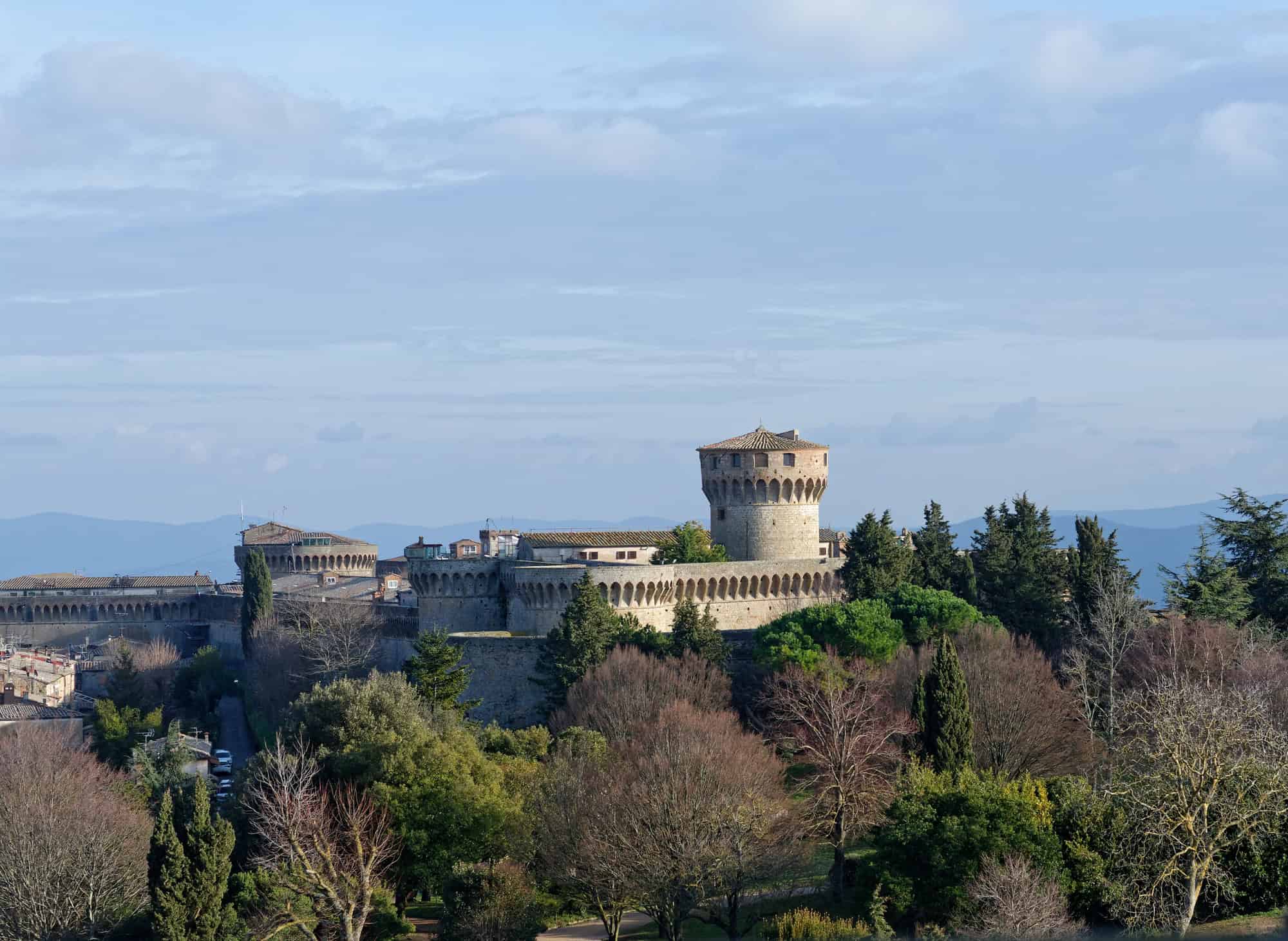 A large stone fortress with round towers stands atop a hill, reminiscent of Tuscany villages, surrounded by trees under a partly cloudy sky.