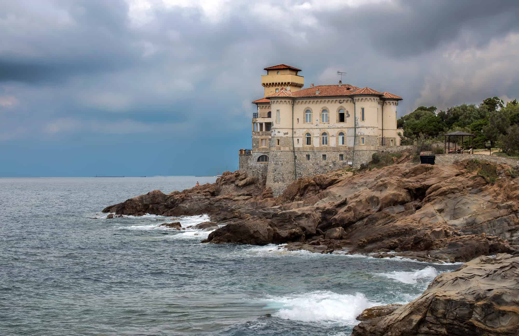 A coastal castle sits on rocky cliffs above the sea under a cloudy sky, evoking the dramatic beauty often found in a Tuscany travel guide.