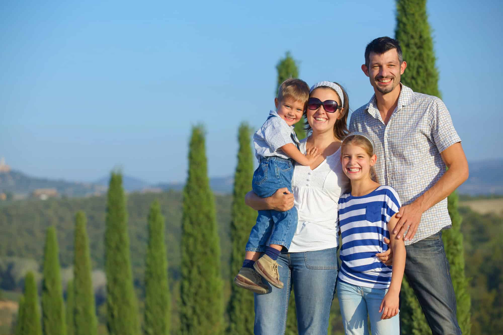 A smiling family of four poses outdoors with trees, hills, and the charming scenery of Tuscany villages in the background.