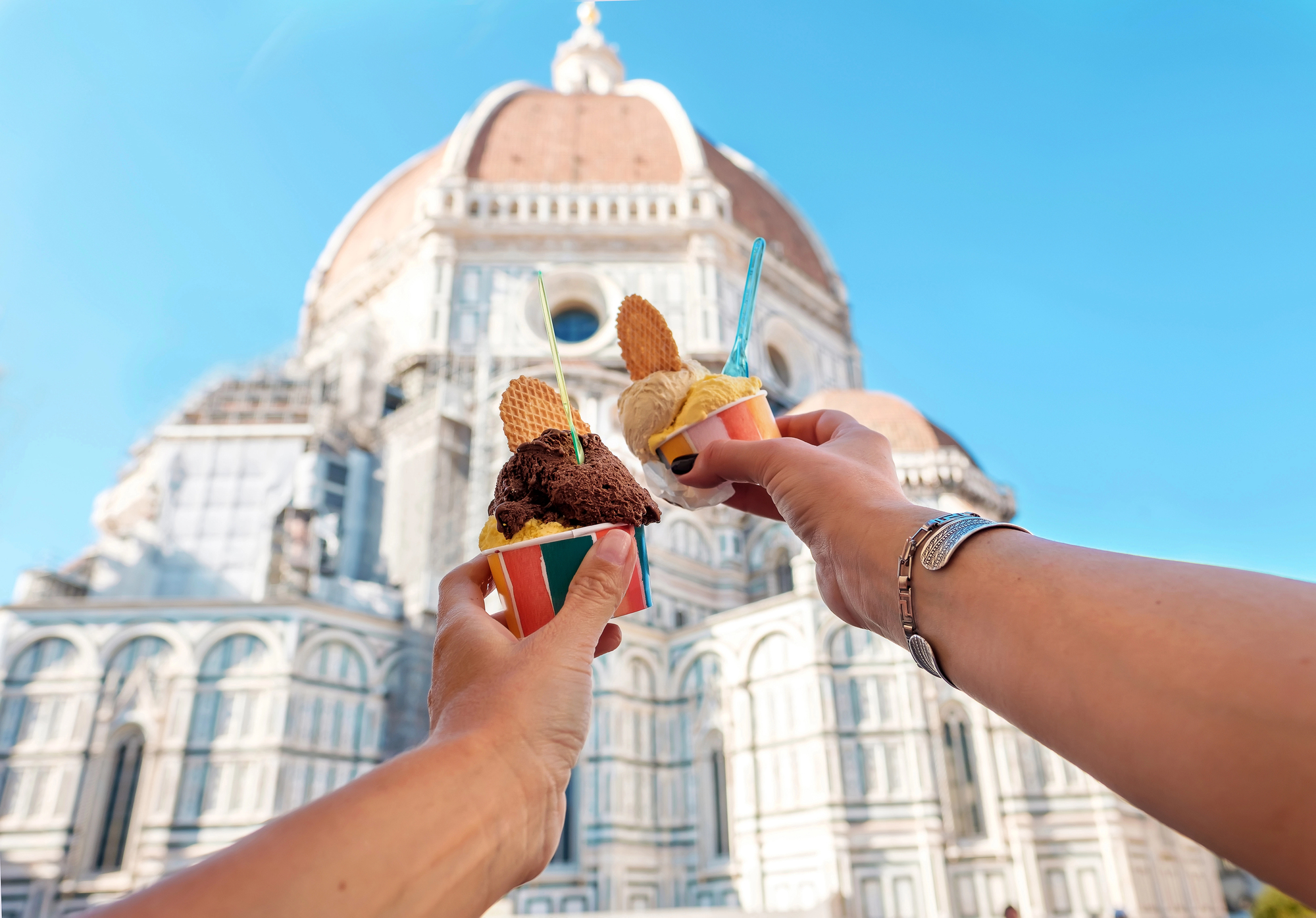 Two hands holding gelato cups with wafers in front of Florence's Cathedral under a bright blue sky, capturing a classic Florence foods moment.