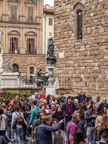 A large crowd gathers near statues and historic buildings in a picturesque Tuscan city square, capturing the timeless charm found in any Tuscany travel guide.
