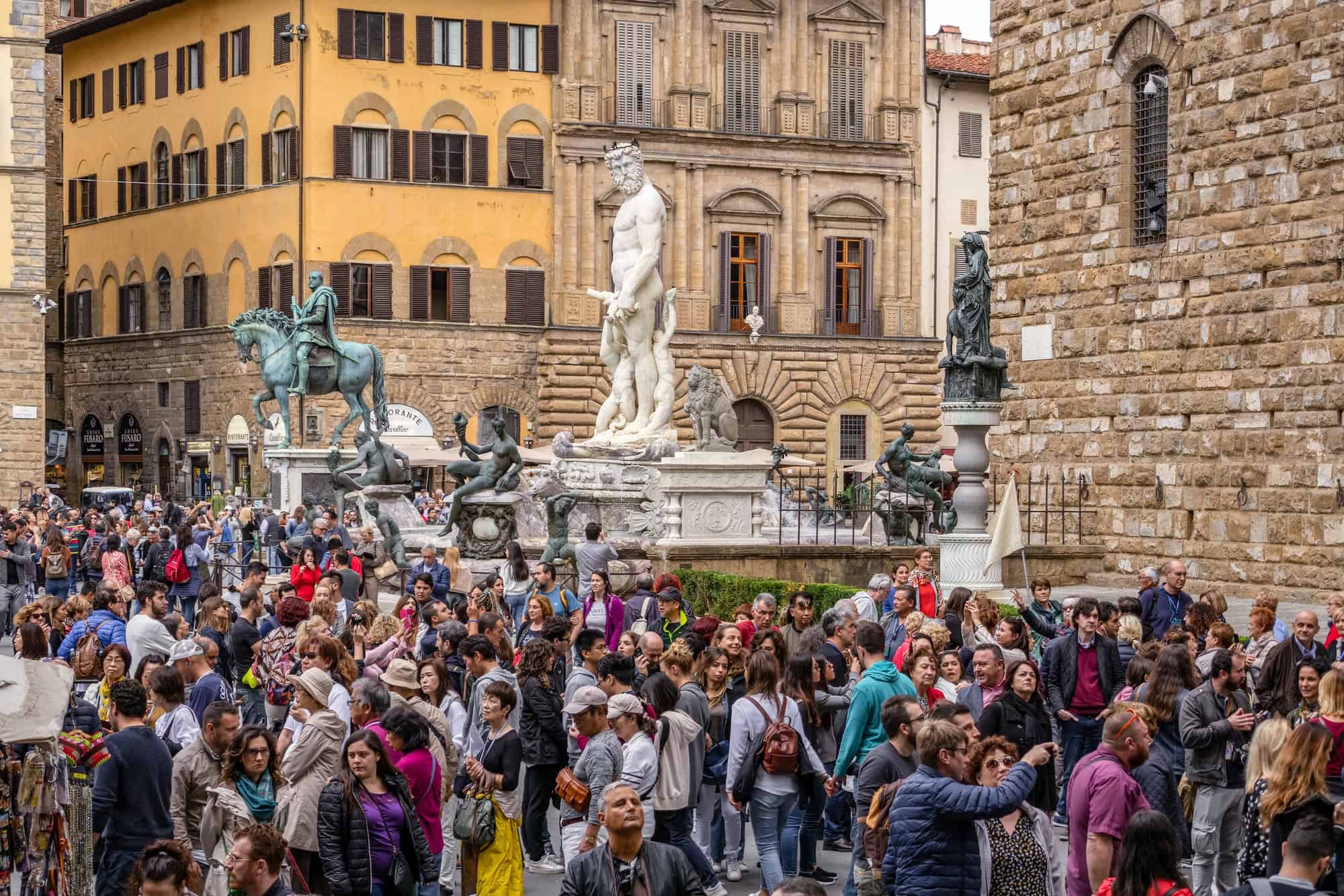 A large crowd gathers near statues and historic buildings in a picturesque Tuscan city square, capturing the timeless charm found in any Tuscany travel guide.
