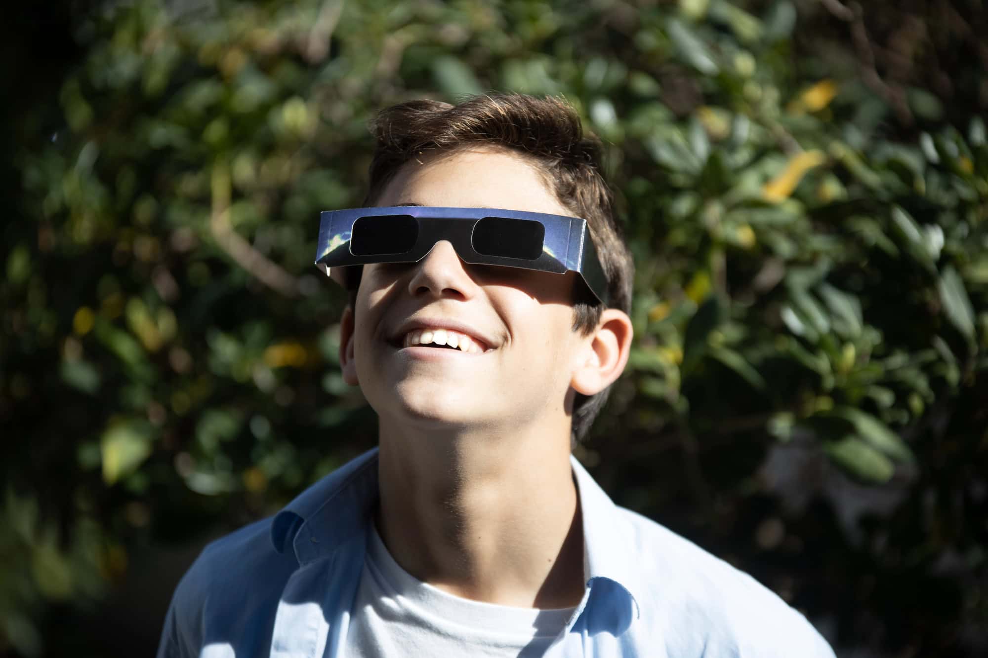 Smiling boy wearing eclipse glasses looks up outside with greenery in the background, dreaming of the Best Places to Catch the 2026 Total Solar Eclipse in Spain and Iceland.
