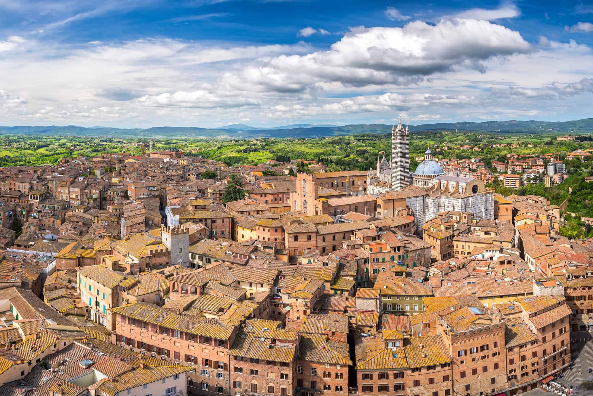 Aerial view of Siena, Italy, with historic buildings, cathedral, and green hills under a partly cloudy sky—an iconic gem among Tuscany villages.