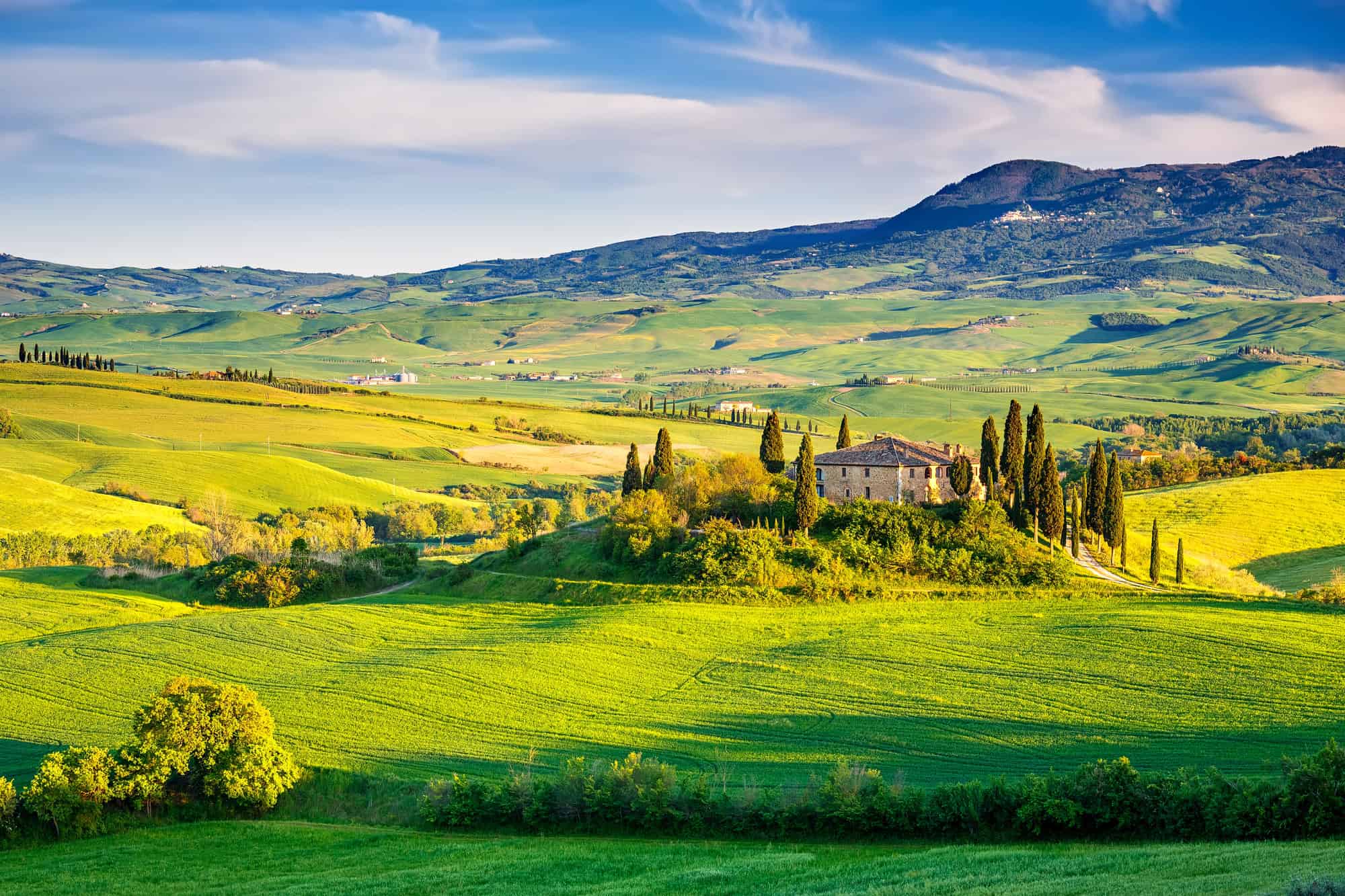 Stone farmhouse surrounded by cypress trees in the rolling green hills of Tuscany under a blue sky with clouds.