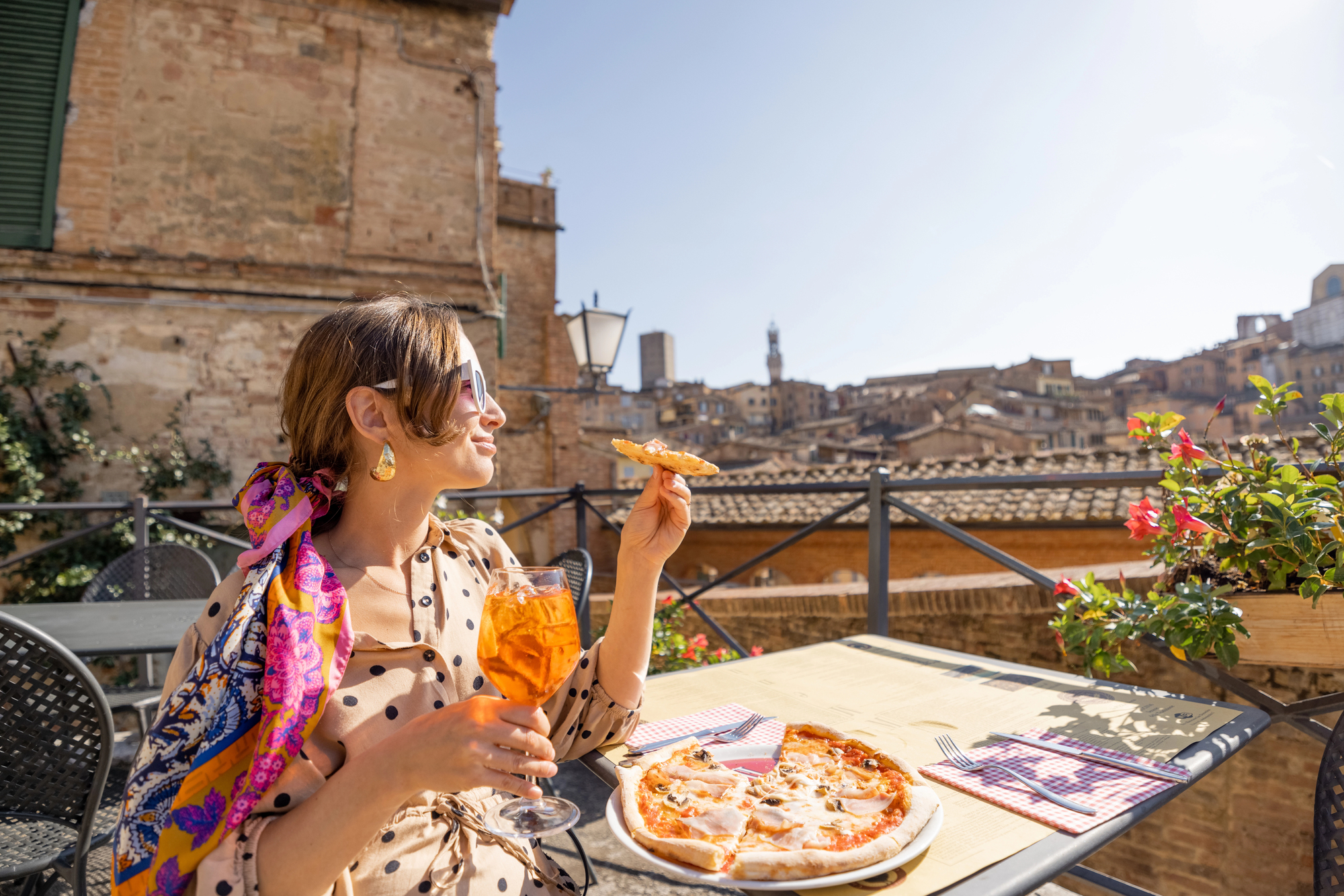 Woman enjoying pizza and an orange drink at an outdoor cafe with a scenic city view inspired by charming Tuscany villages in the background.