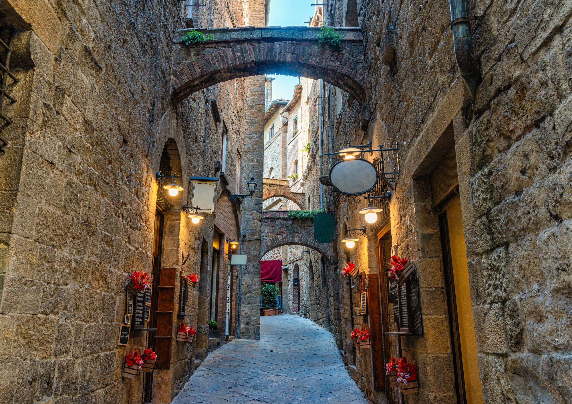 Narrow stone alley with arches, hanging lanterns, and red flowers in pots along the walls, evoking the timeless charm of Tuscany.
