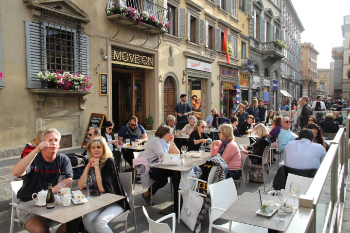 People dining at outdoor tables of a busy caf&eacute; on a sunny day, savoring Florence food in a lively European city street.