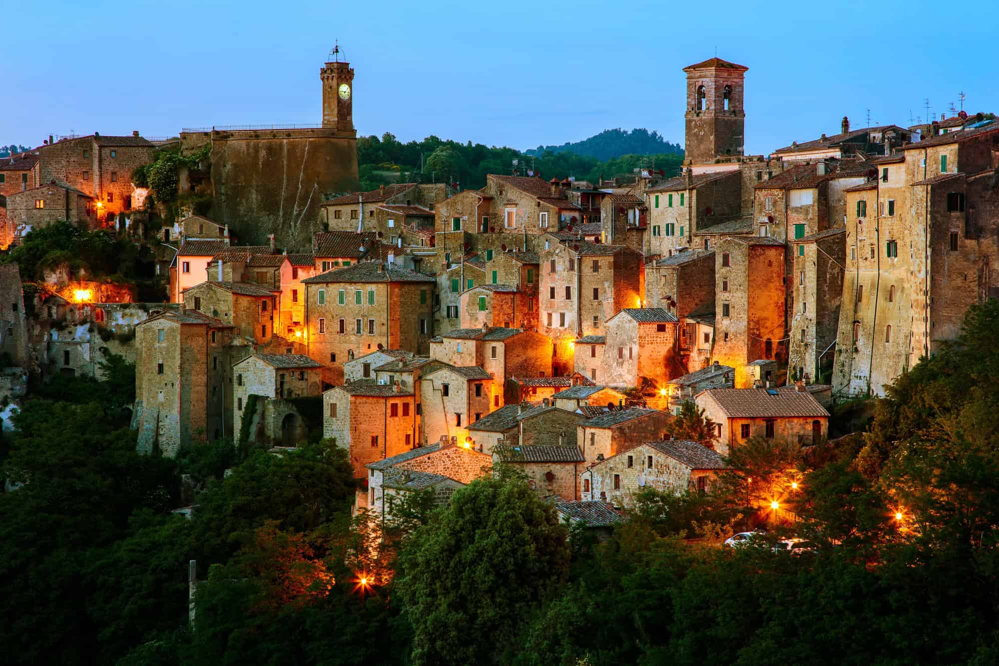 Hilltop village with stone buildings and warm lights glowing at dusk, surrounded by trees and greenery, capturing the timeless charm of Tuscany villages.