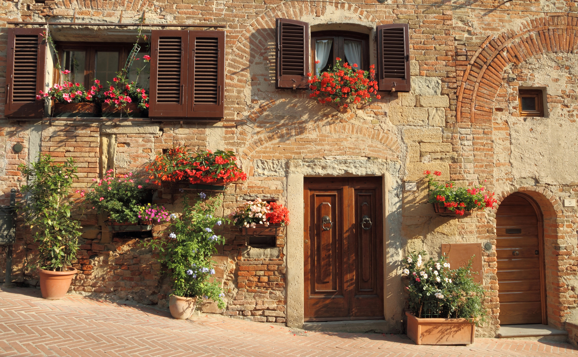 Old brick building with wooden doors, flower boxes, and potted plants on a sunny cobblestone street, reminiscent of charming Tuscany villages.
