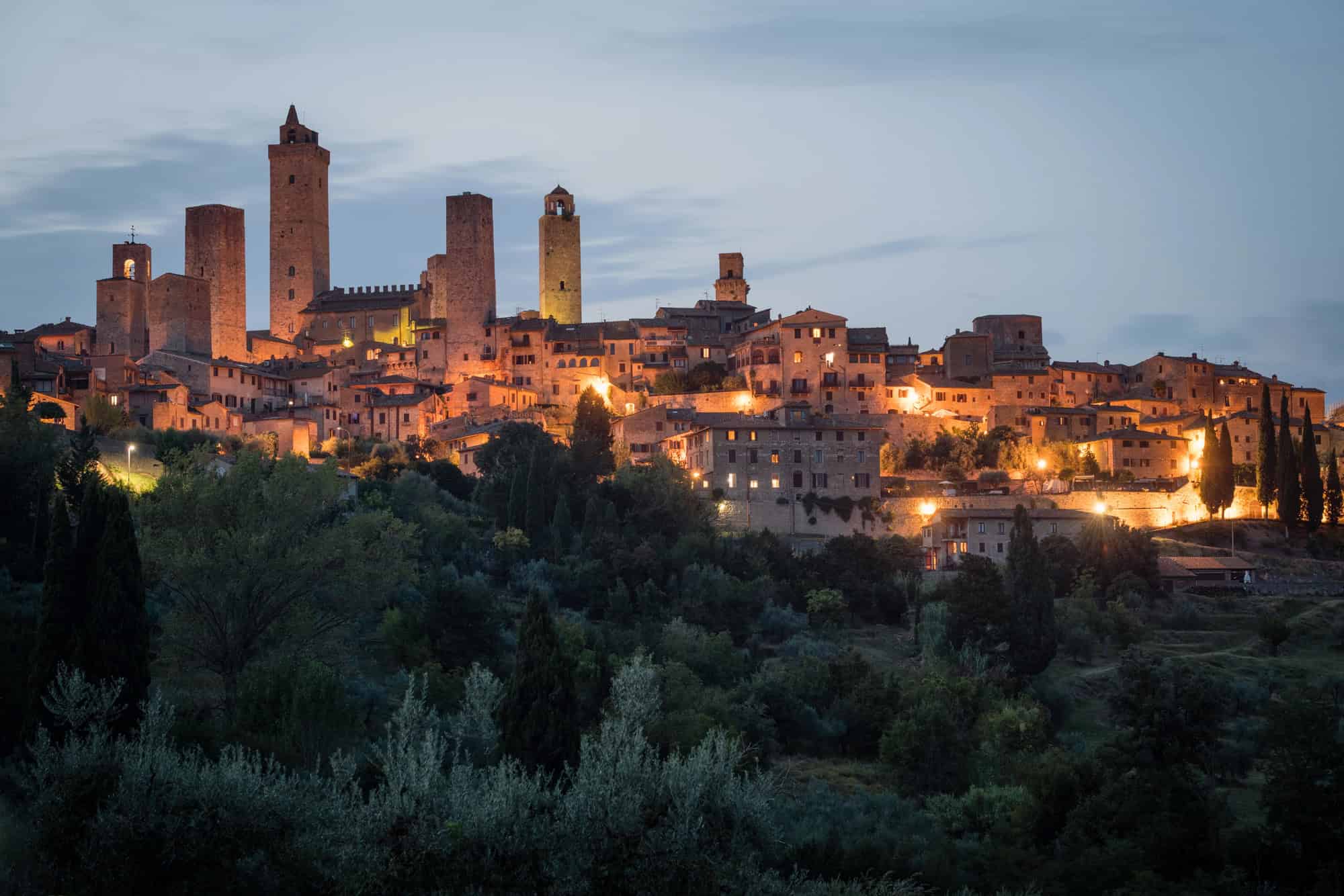 Medieval hilltop town with illuminated stone towers and buildings at dusk, surrounded by greenery—a picturesque gem among Tuscany villages.