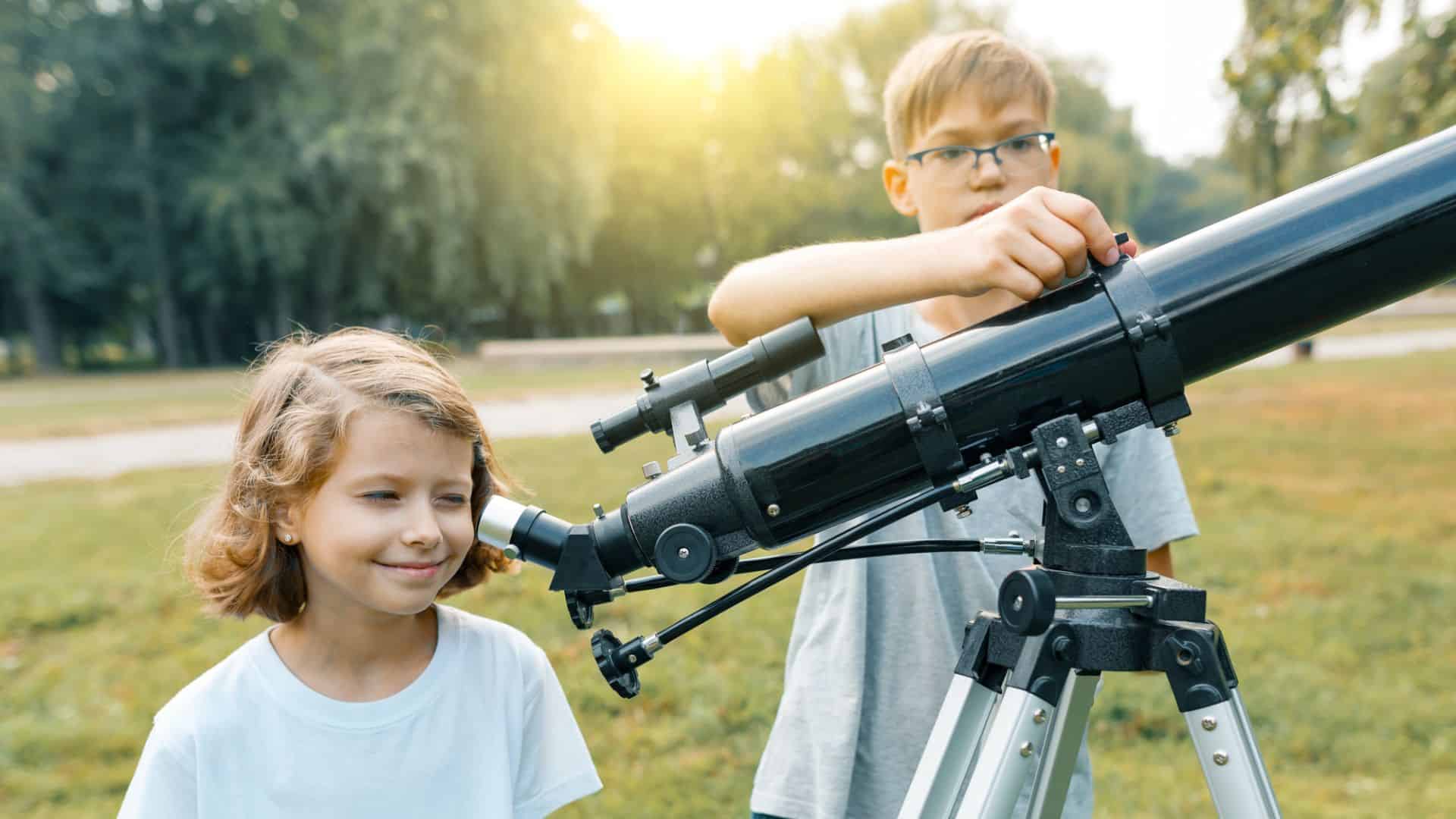 Two children use a telescope outdoors in a park on a sunny day, with trees in the background.