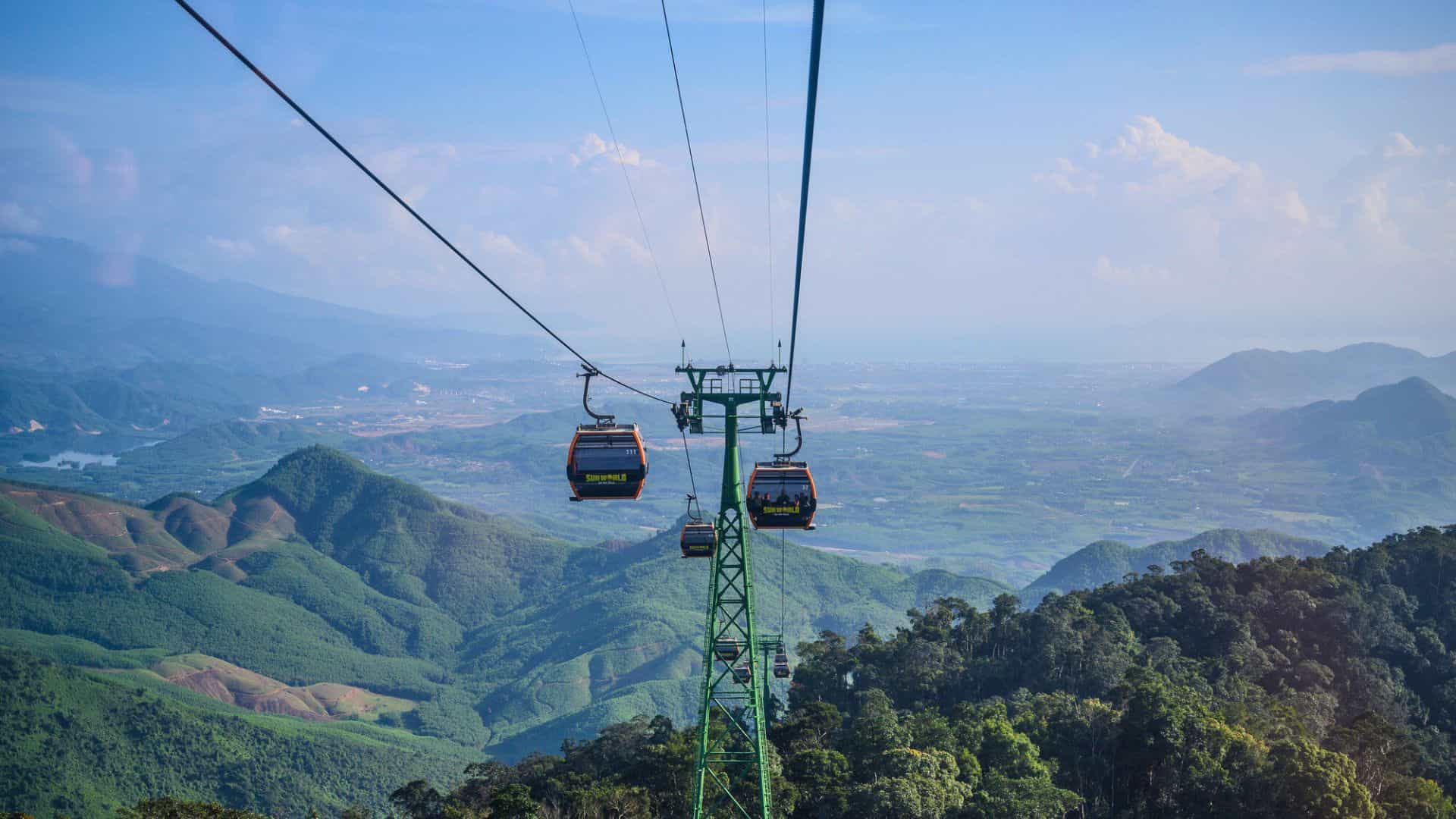 Cable cars travel above lush green mountains, offering a scenic view of the valley and distant blue sky.