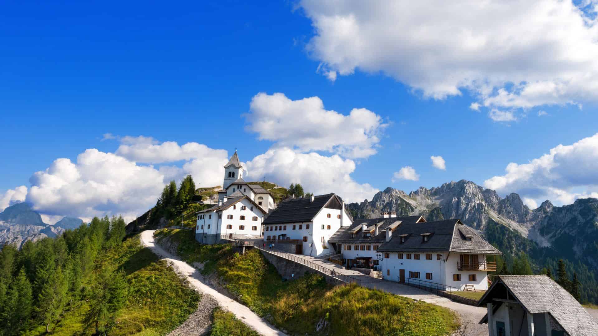 White mountain village buildings with a church, surrounded by green trees and peaks under a blue sky.