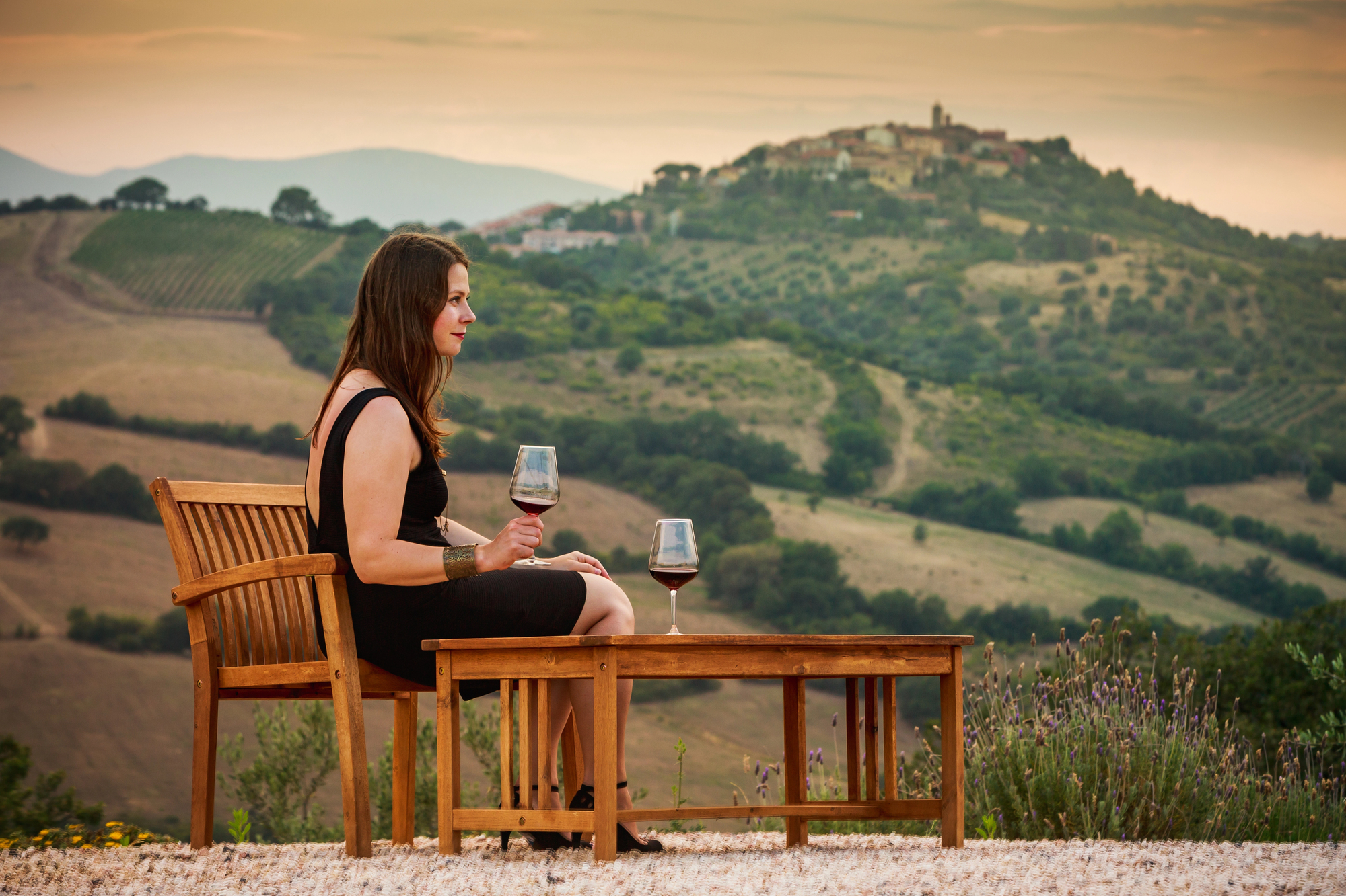Woman in a black dress sits at an outdoor table, holding wine, with rolling hills and a distant village behind her—capturing the elegance of Tuscany wine tours.