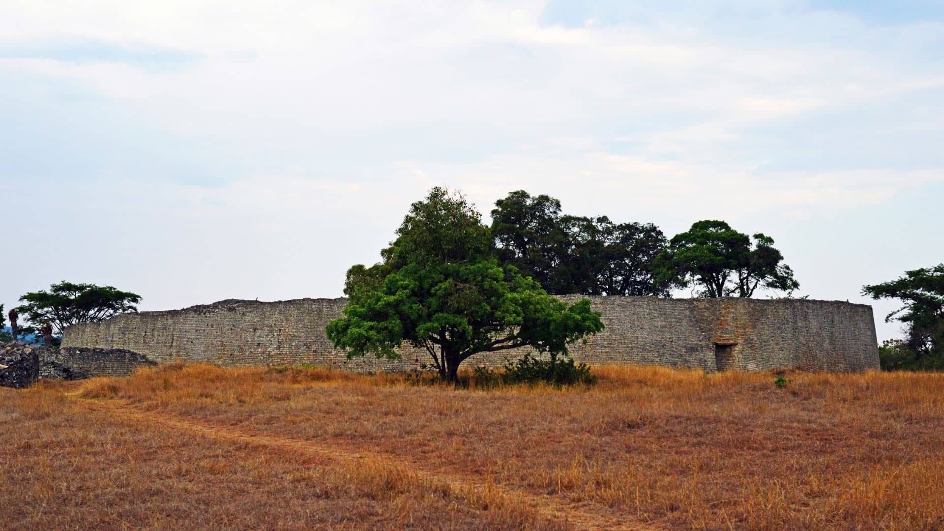 Ancient stone wall structure with green trees and dry grass under a cloudy sky.