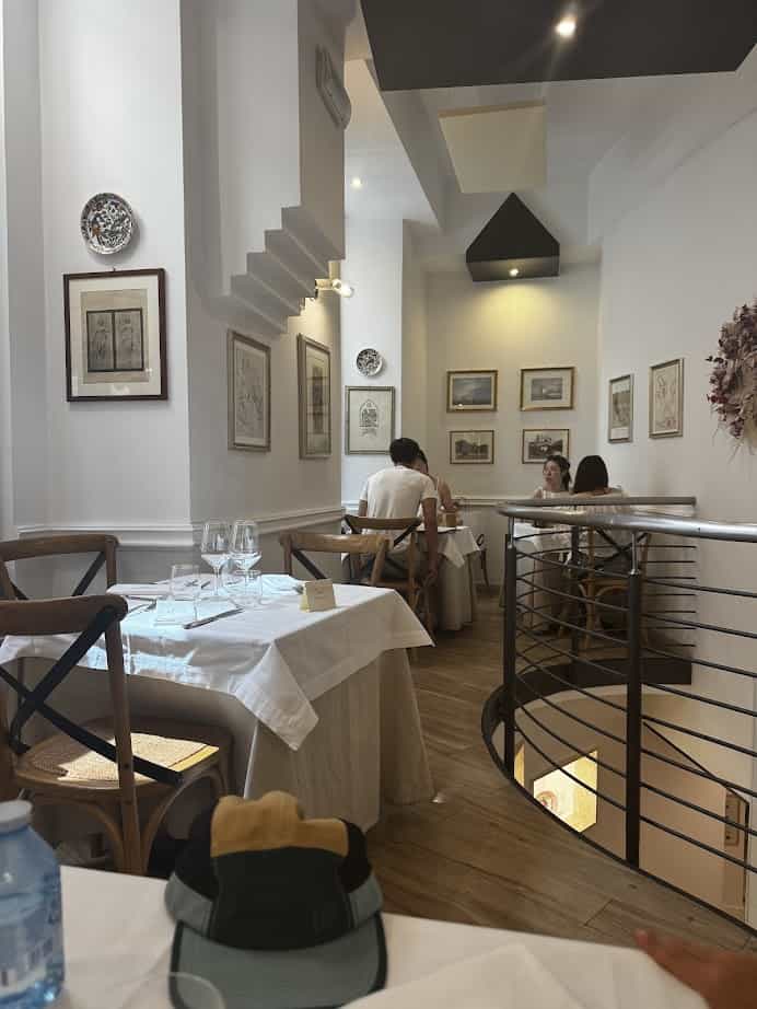 A cozy restaurant interior in Rome with white walls, framed art, set tables, and a spiral staircase in the foreground—perfect for families exploring the city or included in your Family Itinerary.