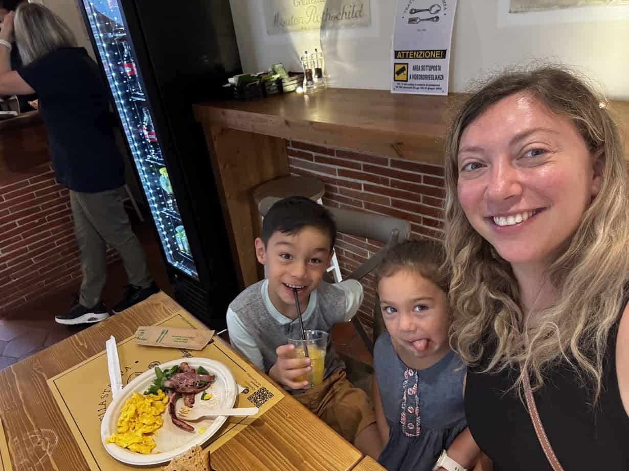 A woman and two kids smile at a restaurant table with food and drinks, enjoying their Rome with Kids adventure as part of their family itinerary.